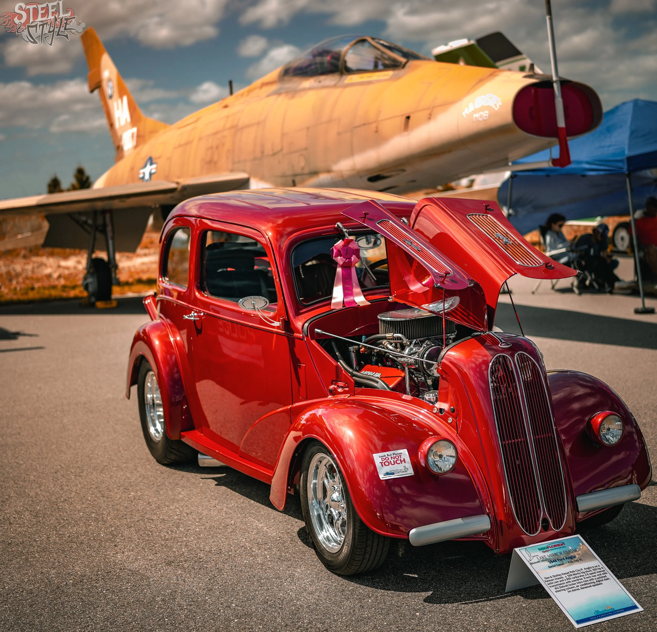 A red vintage car with its hood open, displayed at an outdoor car show, with a yellow military aircraft in the background against a partly cloudy sky.