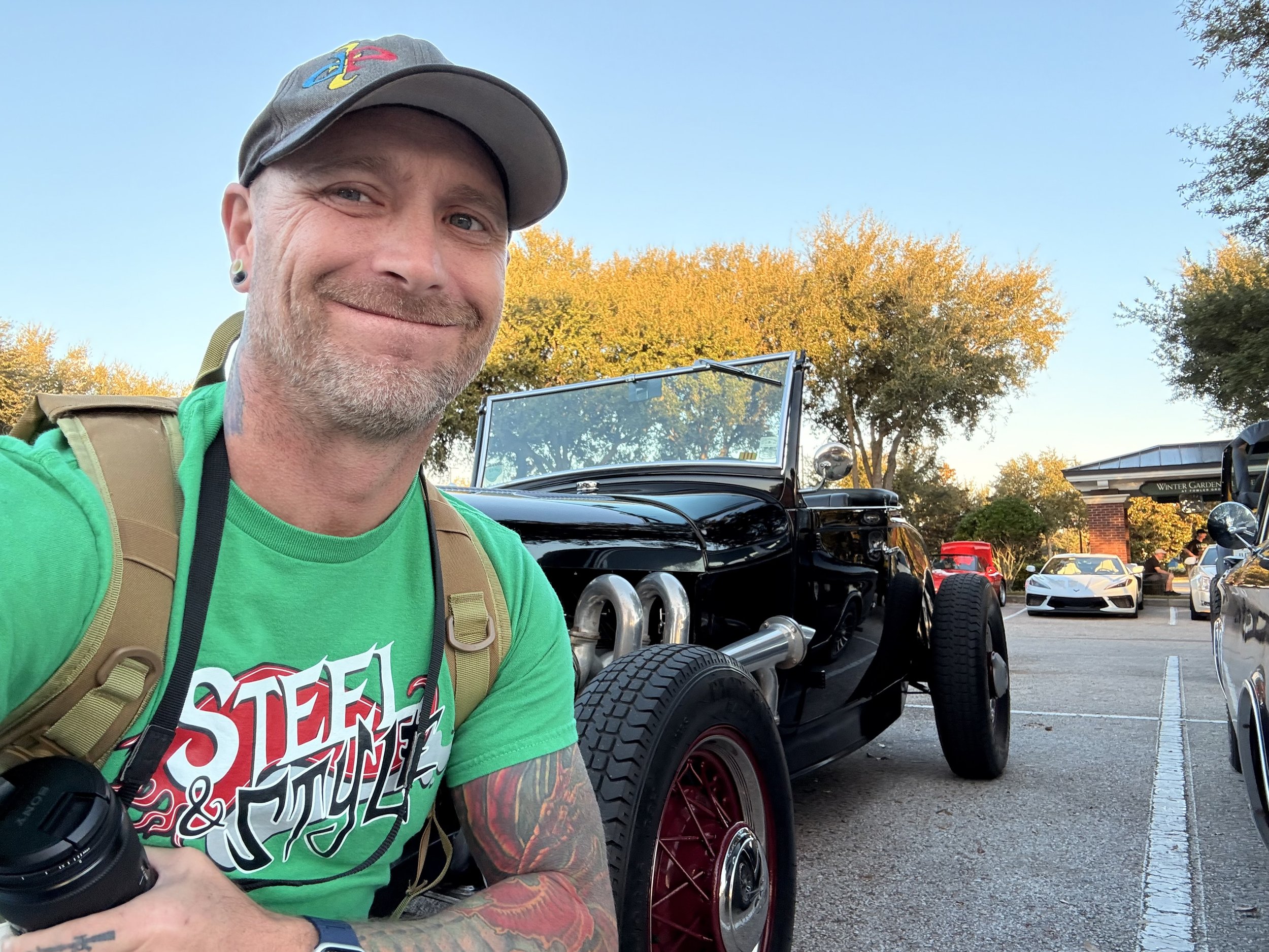 Smiling man with a baseball cap taking a selfie at a car show with classic and sports cars parked behind him.