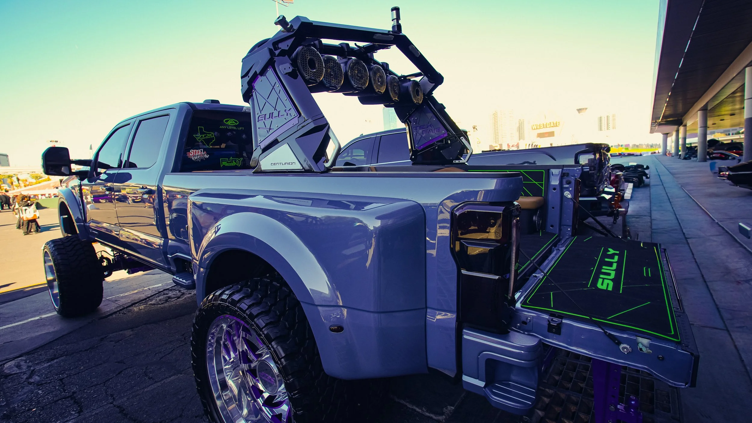 A pickup truck with a lift on the back, equipped with speaker systems and custom modifications, parked outdoors with other vehicles and buildings in the background.