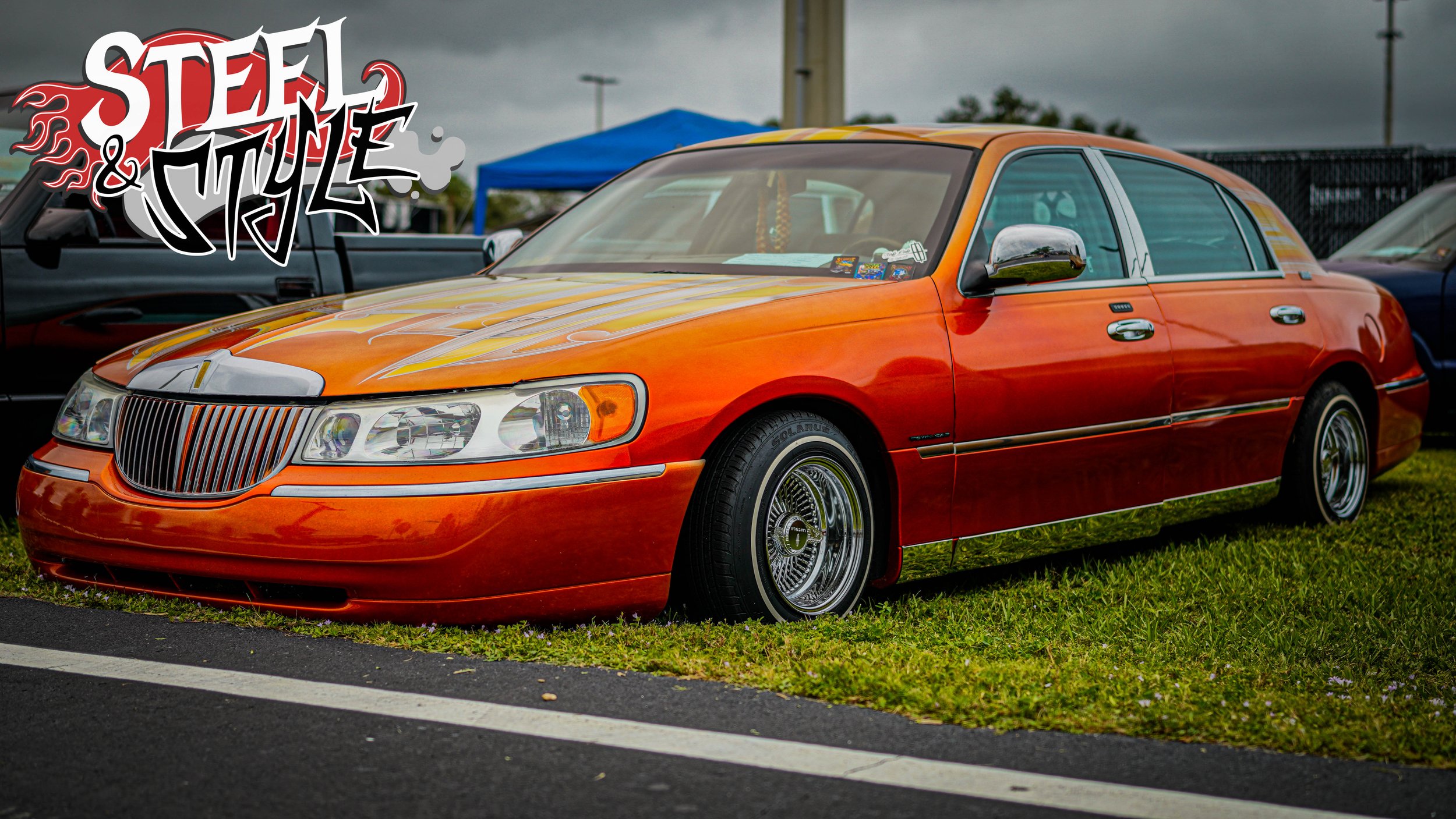 An orange, vintage luxury sedan with shiny chrome accents, parked on grass at a car show, with a graffiti-style ''Steel & Style'' logo overlay in the top left corner.