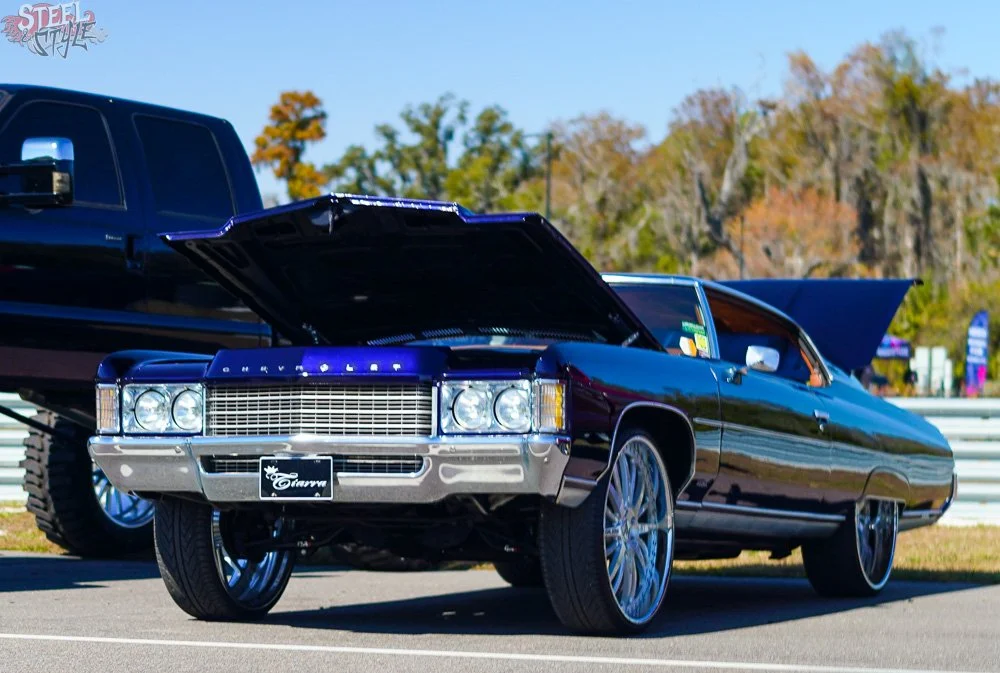 A classic black Cadillac with its hood open, parked outdoors on a sunny day, with trees in the background.