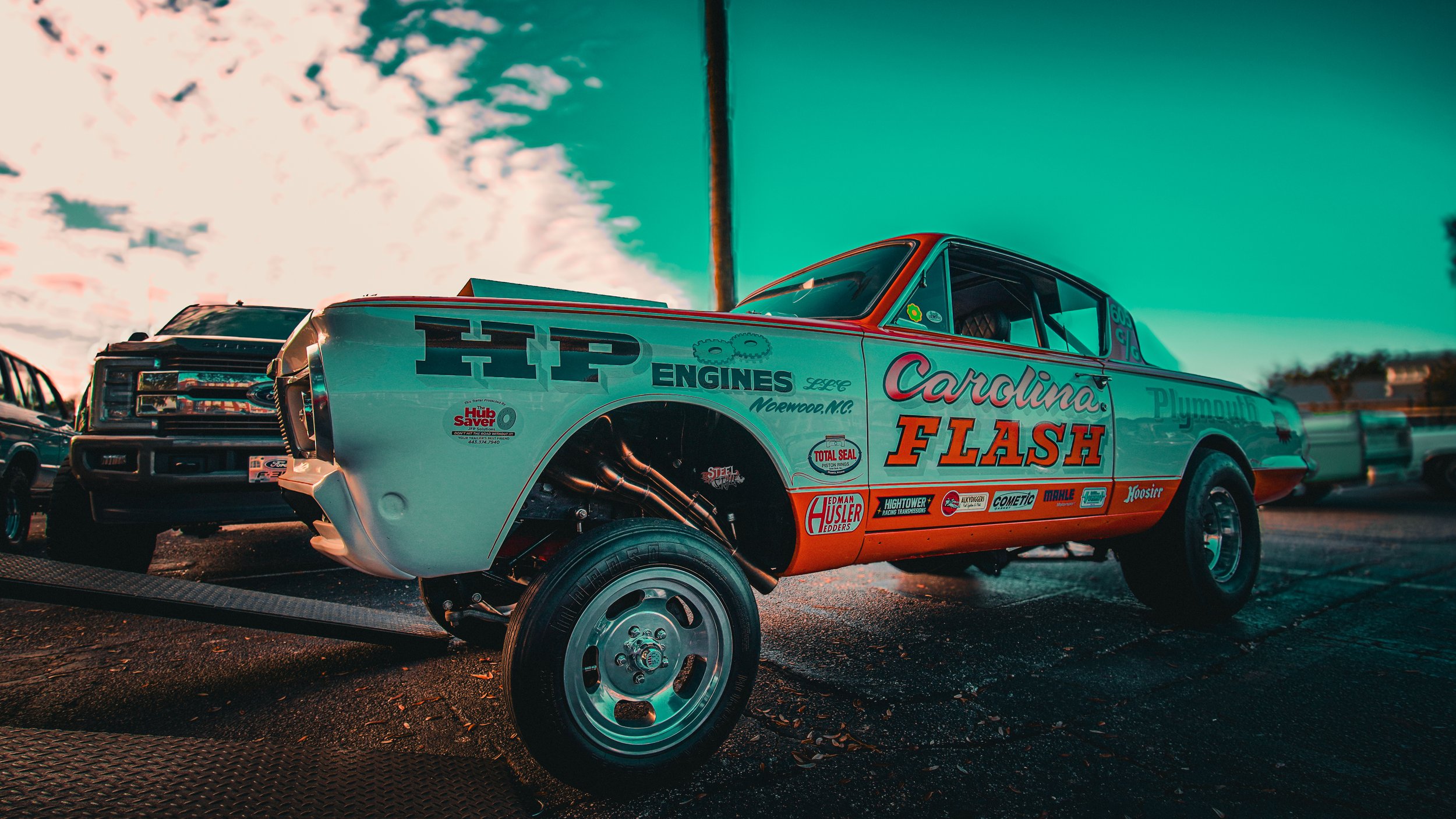 A vintage race car with the words 'Carolina Flash' painted on the side, parked at a race track with other vehicles in the background, under a cloudy sky.