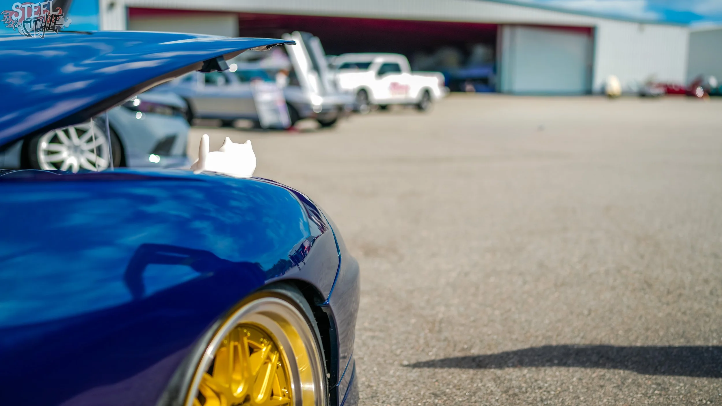 Close-up of the rear side of a blue sports car with yellow wheels parked in an outdoor parking lot, with other vehicles and a storage building in the background.
