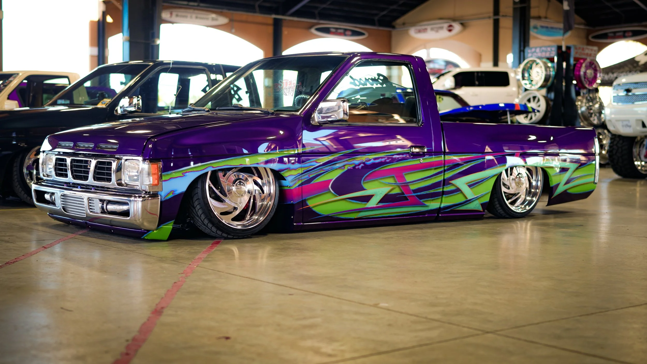 Custom lowrider truck with vibrant purple paint, green and pink graphic design, chrome wheels, and lowered stance, on display indoors at a car show.