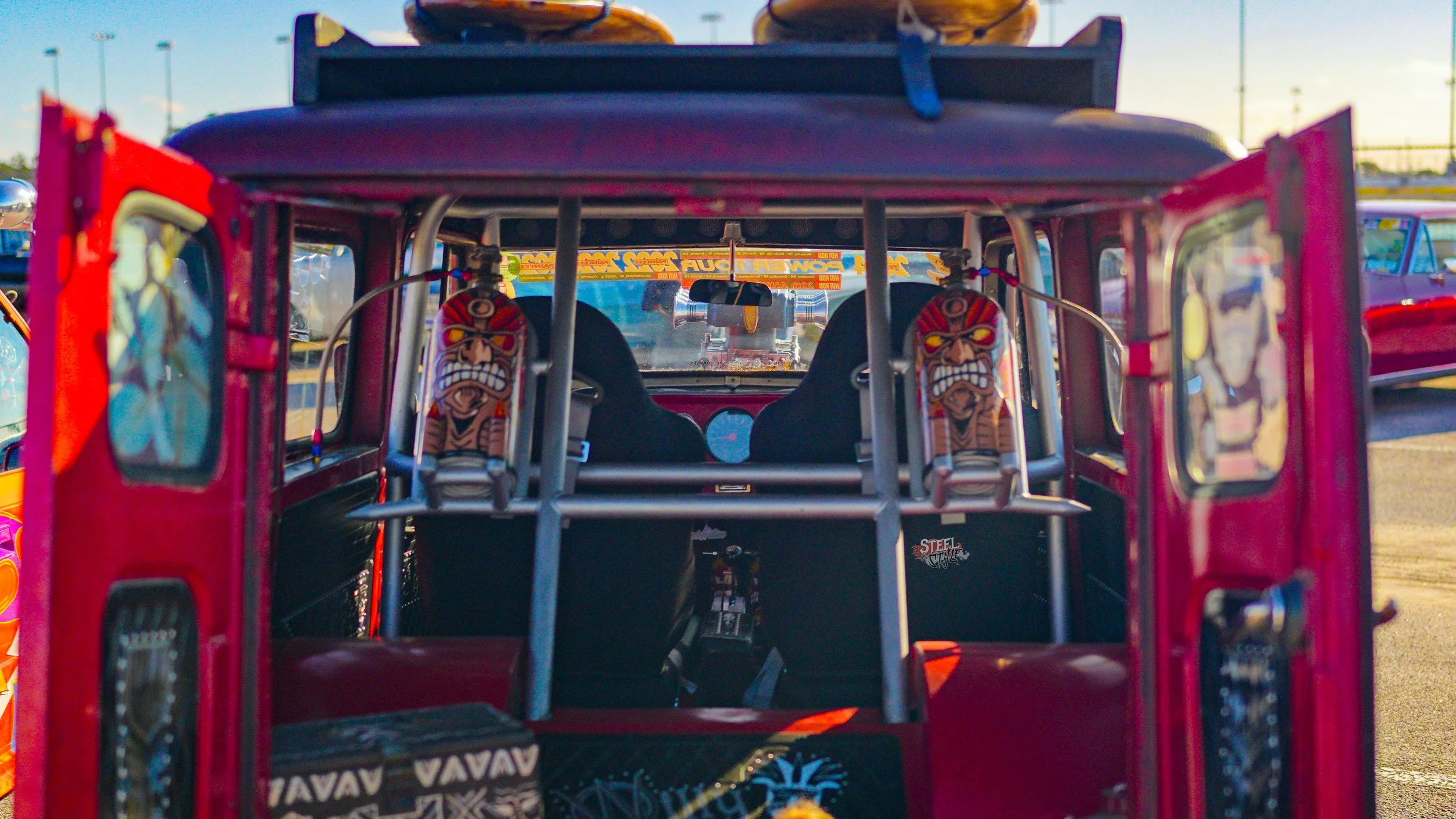 Back of a red go-kart with tribal mask stickers on the seats, inside a parking lot with other cars visible in the background.