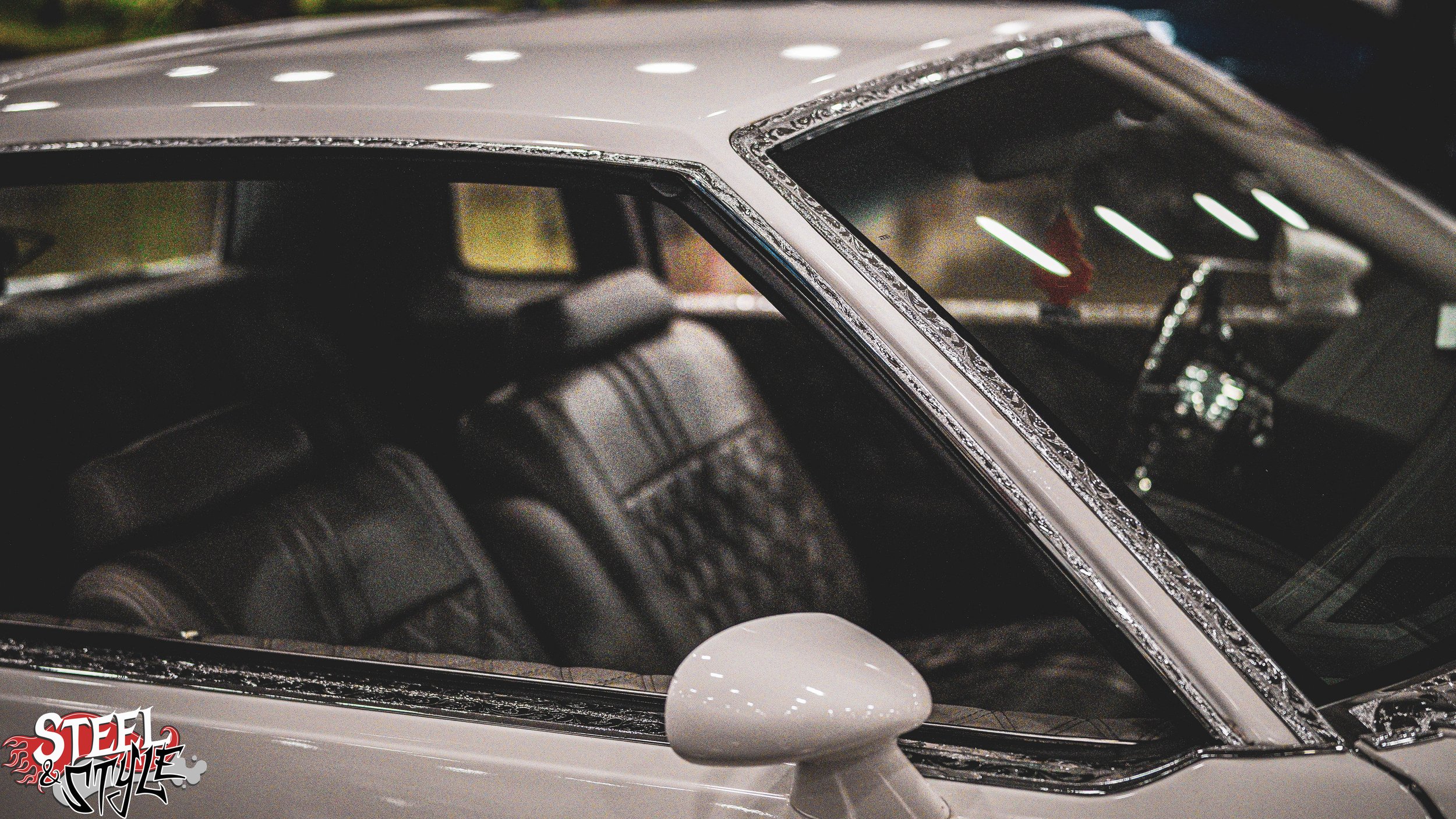 A close-up of a vintage silver car with black leather seats visible through the window, decorated with rhinestones around the windshield, and a side mirror in the foreground. The car has a sleek, classic design.
