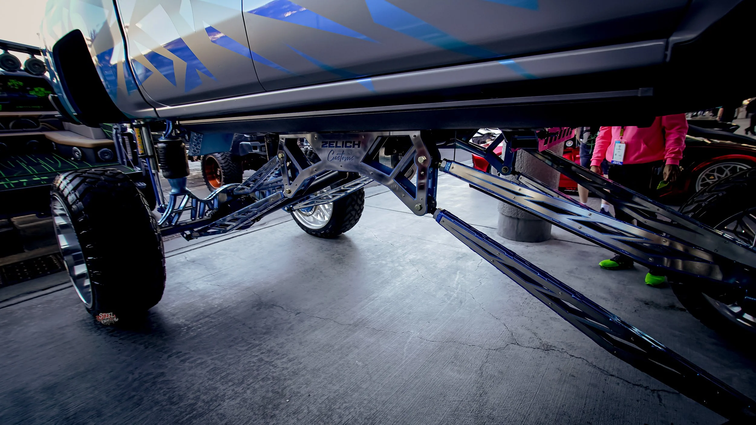 Close-up of a futuristic vehicle chassis on display, featuring metallic components and large tires, with people in the background.