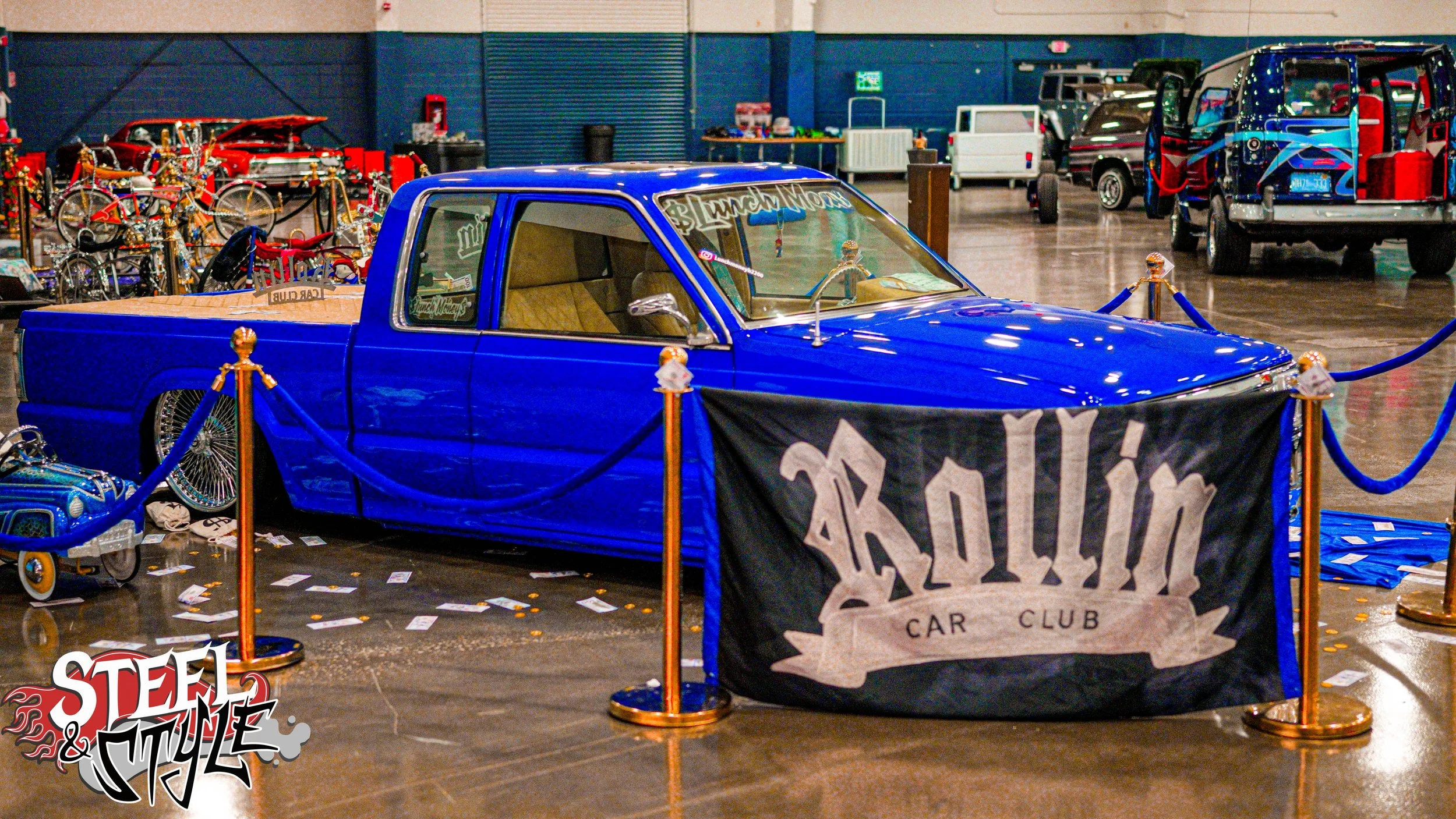A green pickup truck on display at an indoor car show, decorated with a blue paint job and surrounded by blue ropes and a banner reading 'Kowlin Car Club.' In the background, there are other vintage and custom vehicles and bicycle displays.