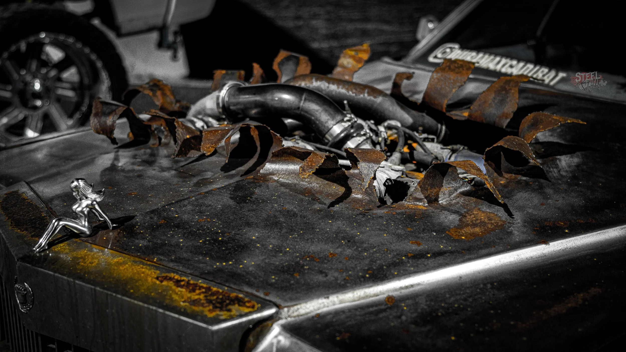 Close-up of a weathered, rusted car hood with two small metallic figurines of women attached; one in a reclining pose on the edge and the other partially visible near rusted machinery.