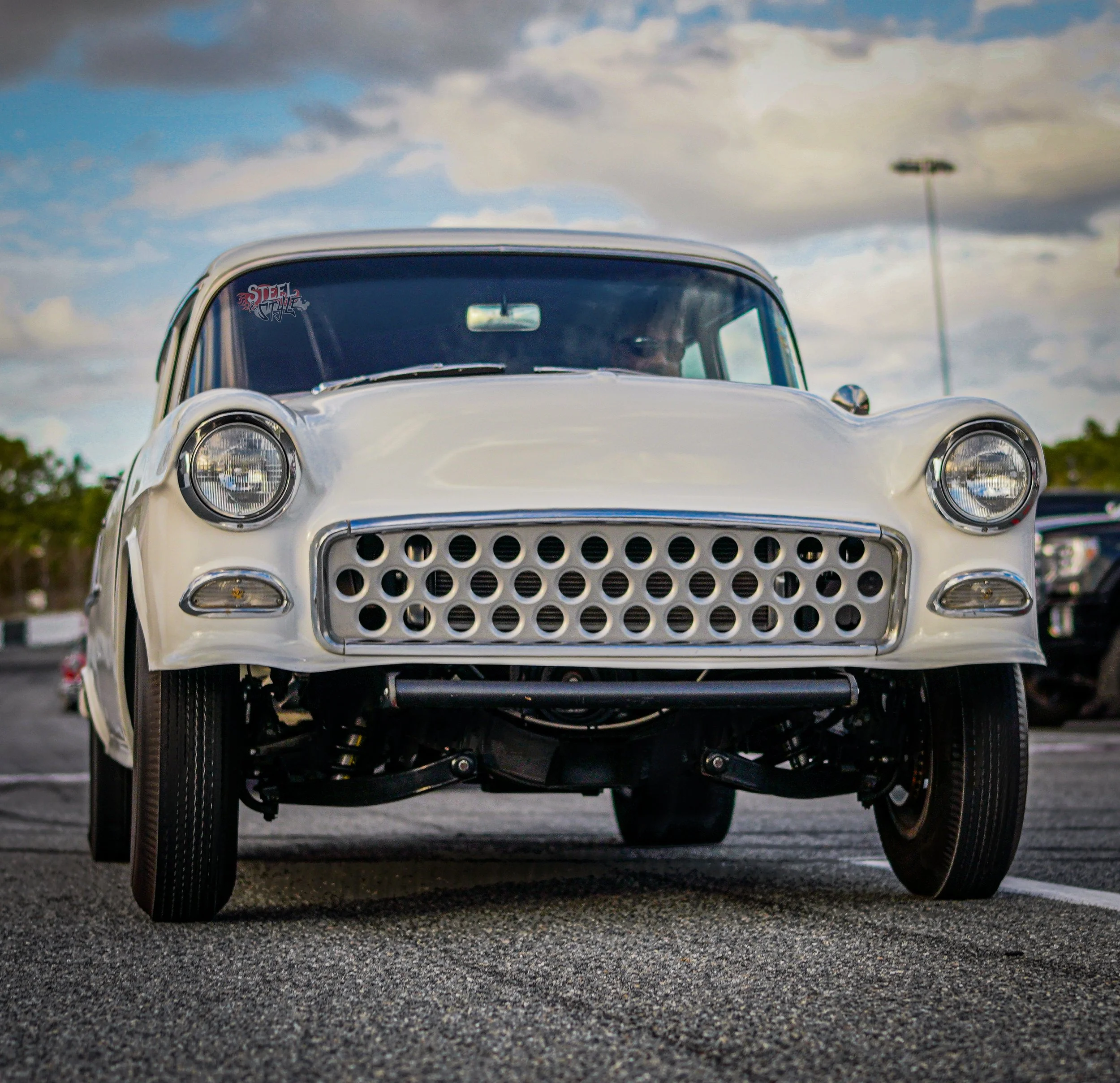 Front view of a vintage white car parked in a lot with a cloudy sky in the background.