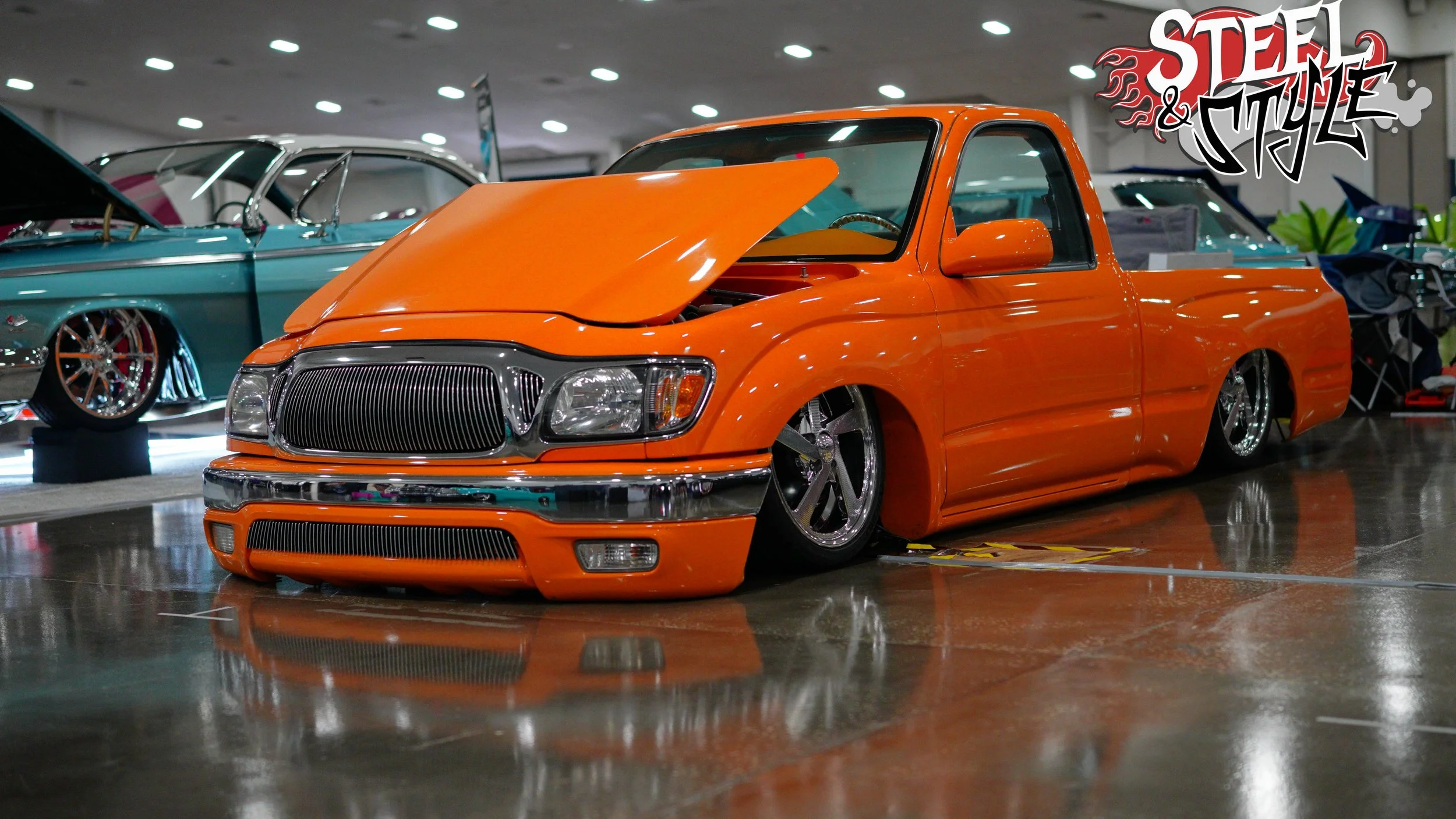 Orange lowrider pickup truck on display at car show, with hood partially open, lowered suspension, and chrome wheels, with other cars in the background.