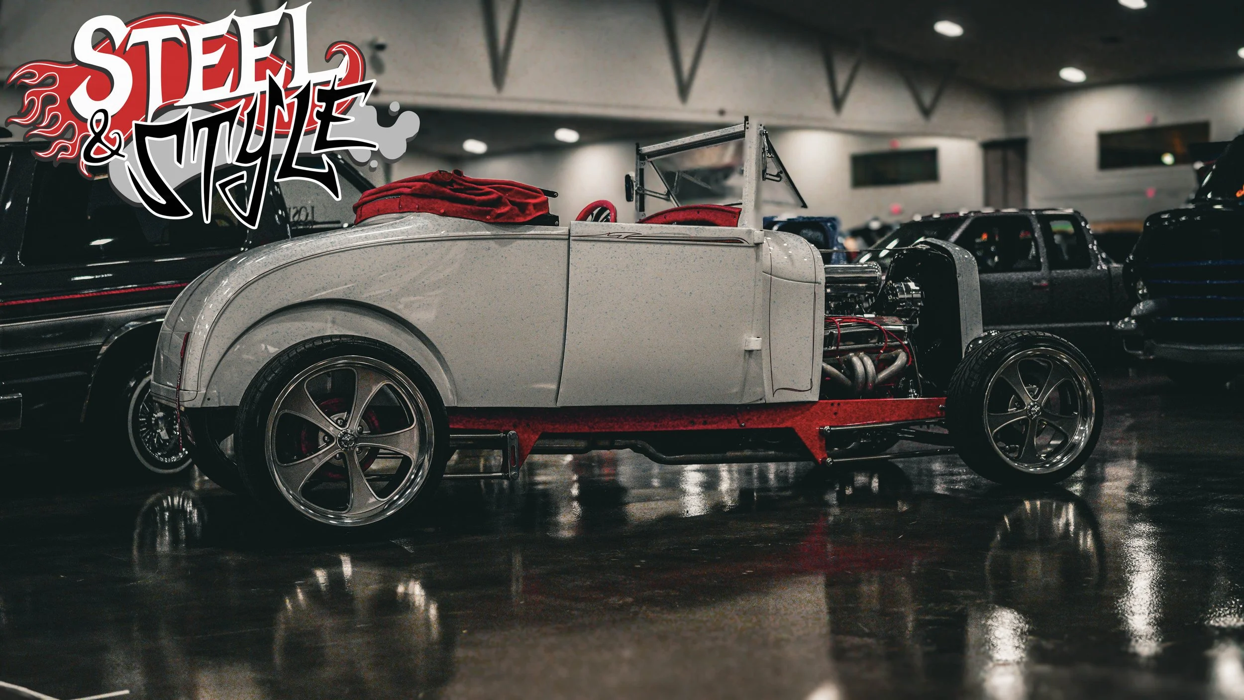 Vintage car with exposed engine on display at an indoor car show, with a 'Steel & Style' logo in the top left corner.