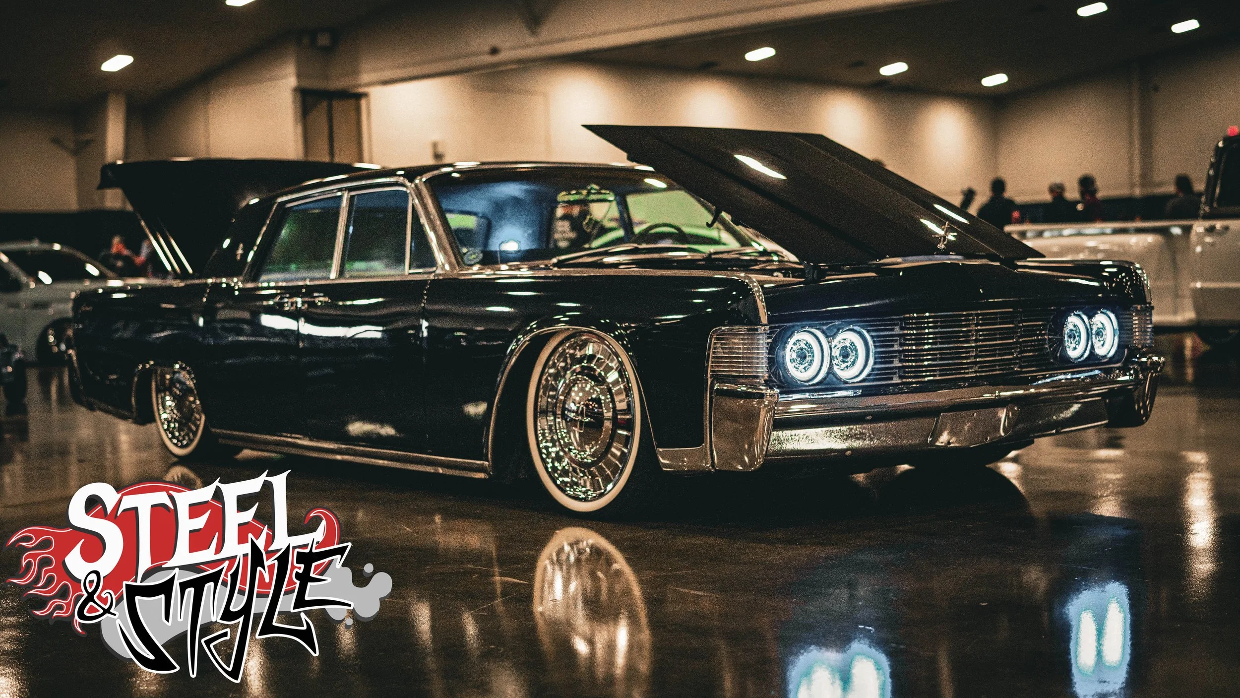 A vintage black Cadillac with its hood open, displayed at a car show titled 'Steel & Style' with other cars and people in the background.