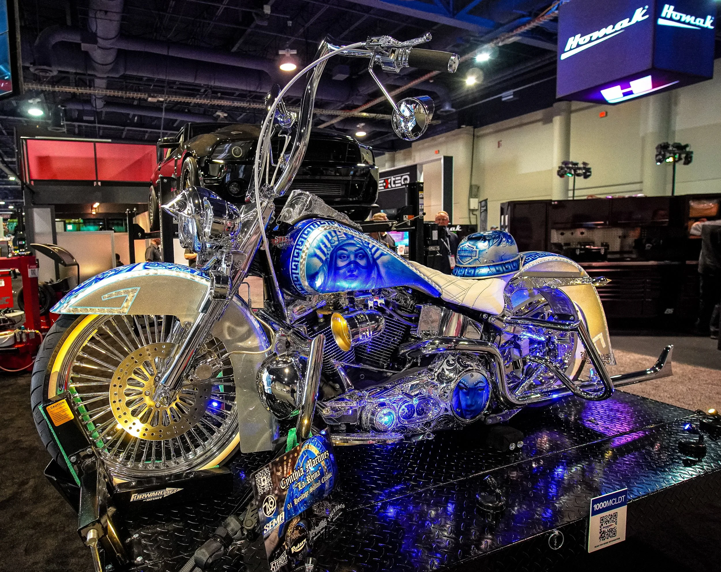 A custom motorcycle decorated with blue and gold accents and illuminated with blue lights, displayed at an indoor exhibition.