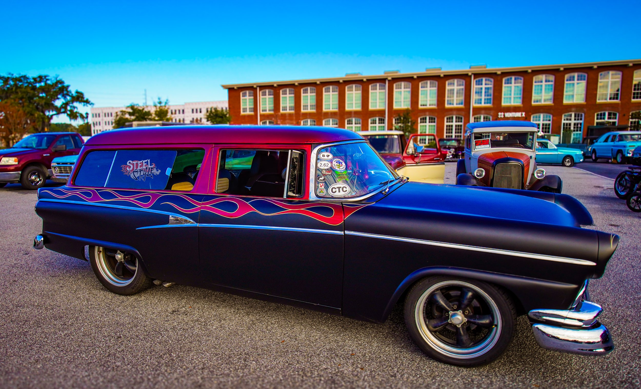 A vintage black station wagon with purple and yellow flames parked at a car show, surrounded by other classic cars, with a brick building and clear blue sky in the background.