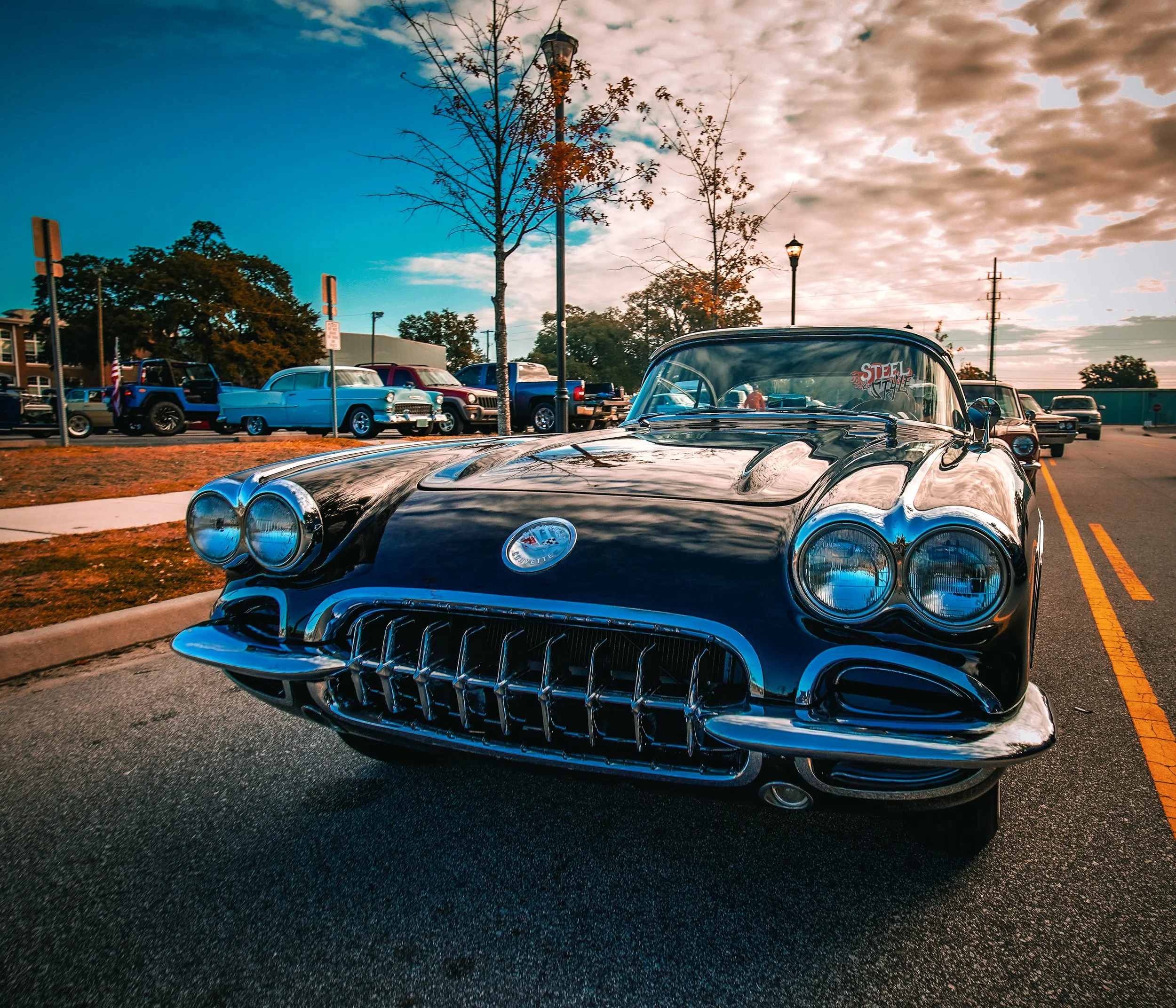 A black vintage Chevrolet Corvette parked on the street during sunset, with other classic cars in the background.