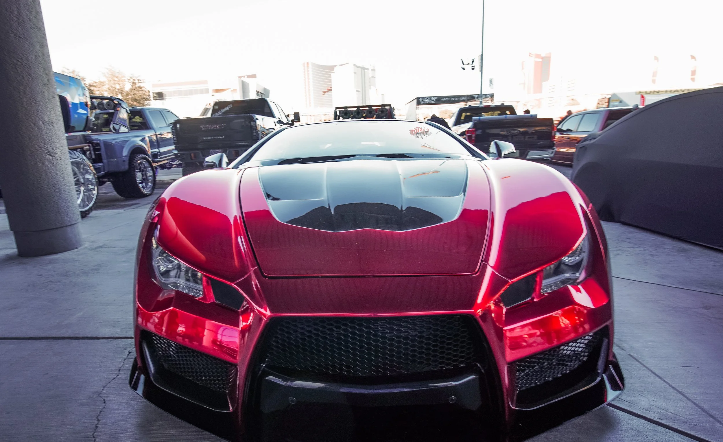 A red sports car with a black hood and windshield, displayed at a car show with other vehicles in the background.