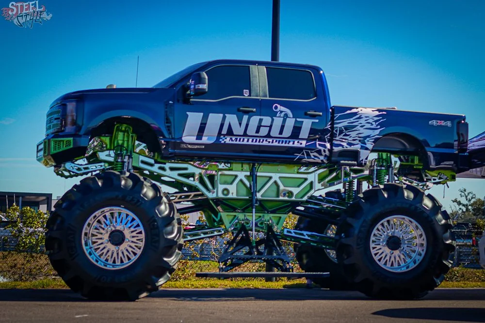 A large blue monster truck with oversized tires and green suspension components, displaying the logo 'UNCUT Motorsports' on the side, parked outdoors against a clear blue sky.