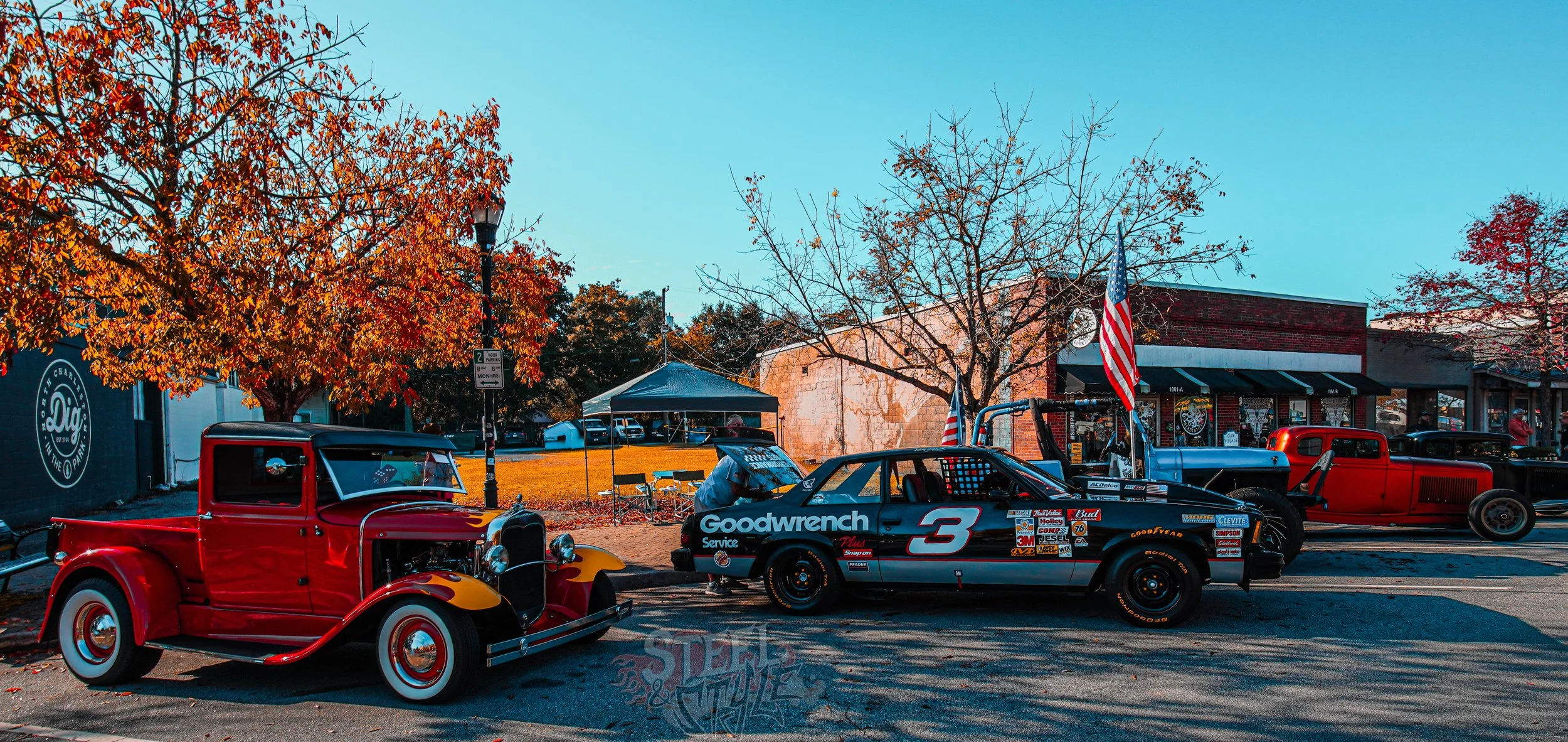 A classic red pickup truck and a vintage race car with sponsorship decals parked on a street during fall with orange and red leaves on trees, and buildings in the background.