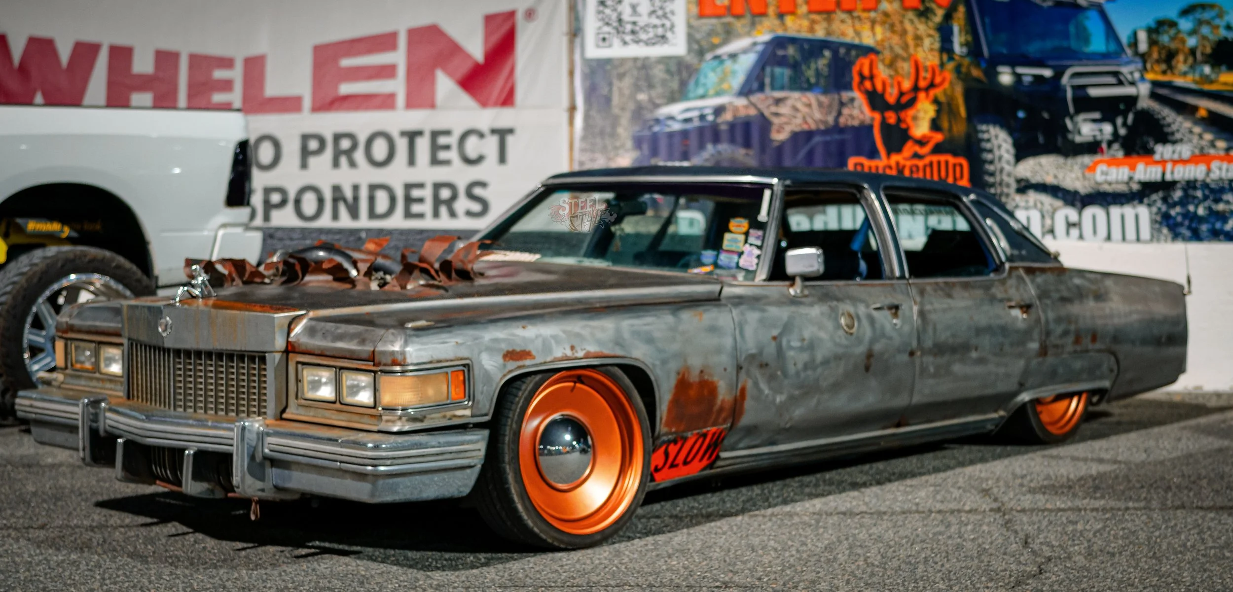 A vintage lowrider car with a weathered, rusted metal body and bright orange wheels, parked outdoors near a truck and a colorful mural.