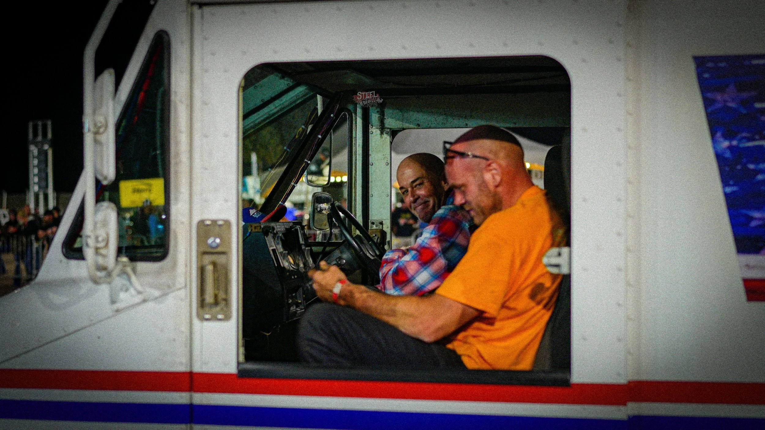 Two men sitting in the cockpit of a race car, smiling and talking with each other at night, with a crowd and lights visible outside.