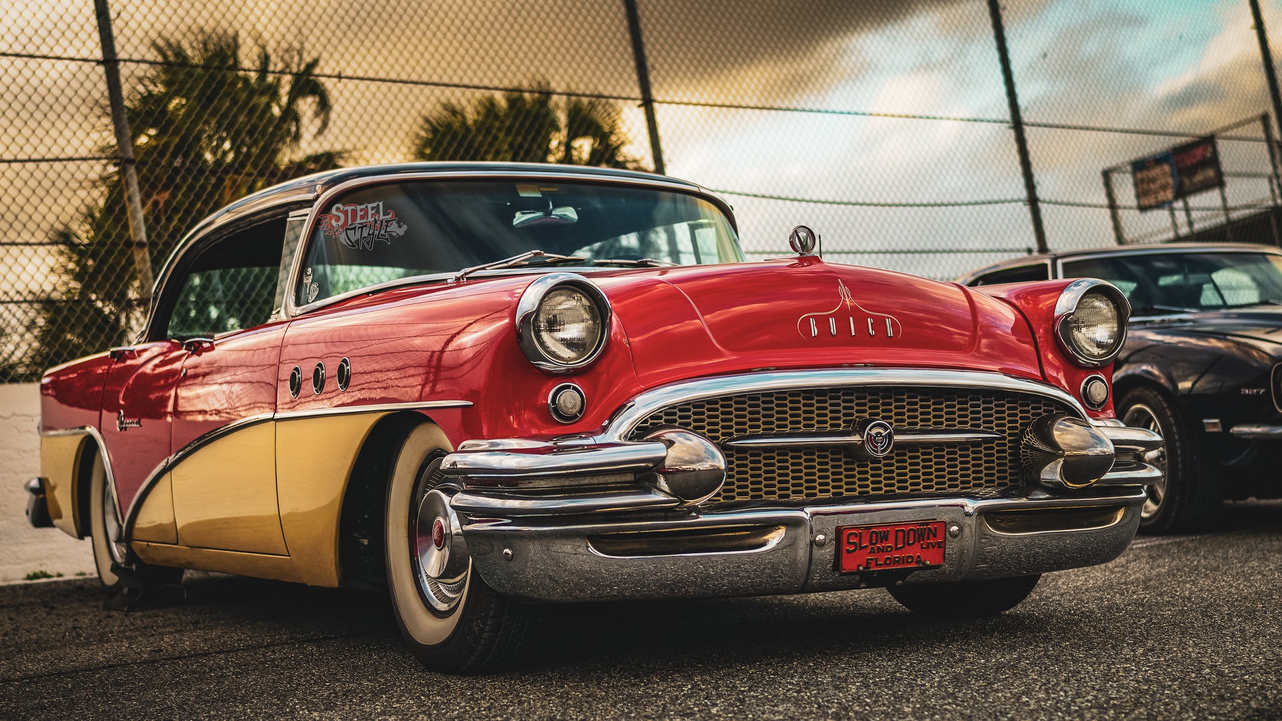 A vintage red and yellow Buick car parked on a lot with a chain-link fence in the background and cloudy sky.
