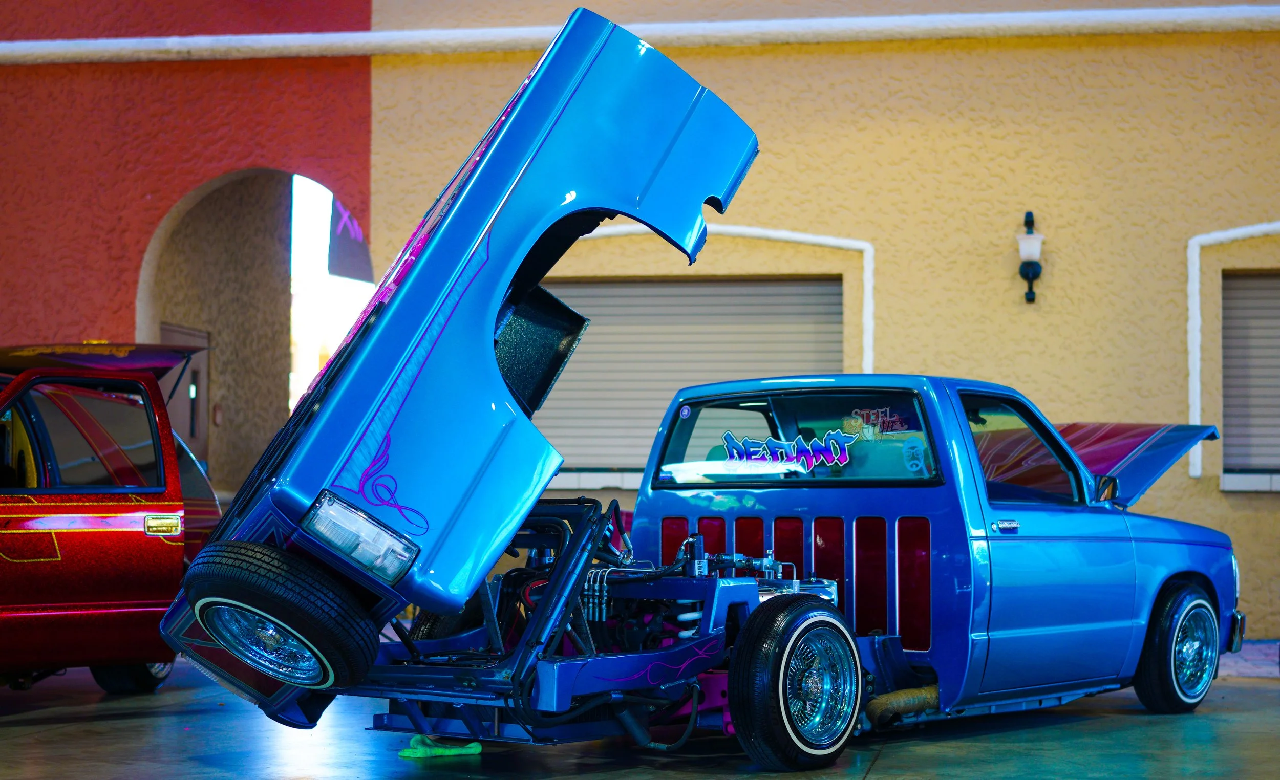 A brightly colored, modified pickup truck with a raised hood and exposed engine, parked indoors with a yellow building in the background.