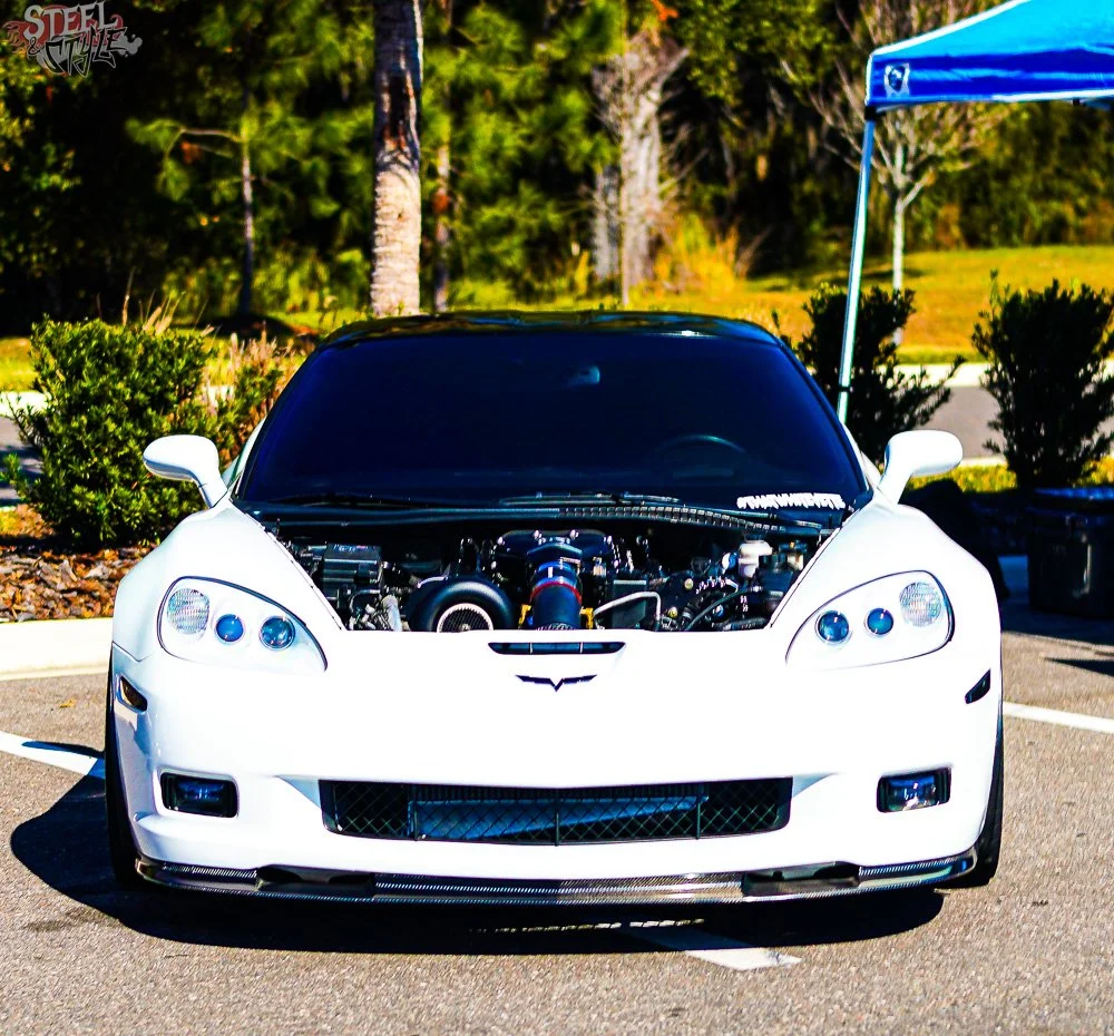 White sports car with a black hood, exposed engine, parked in a lot with trees and bushes in the background, and a blue canopy on the right.