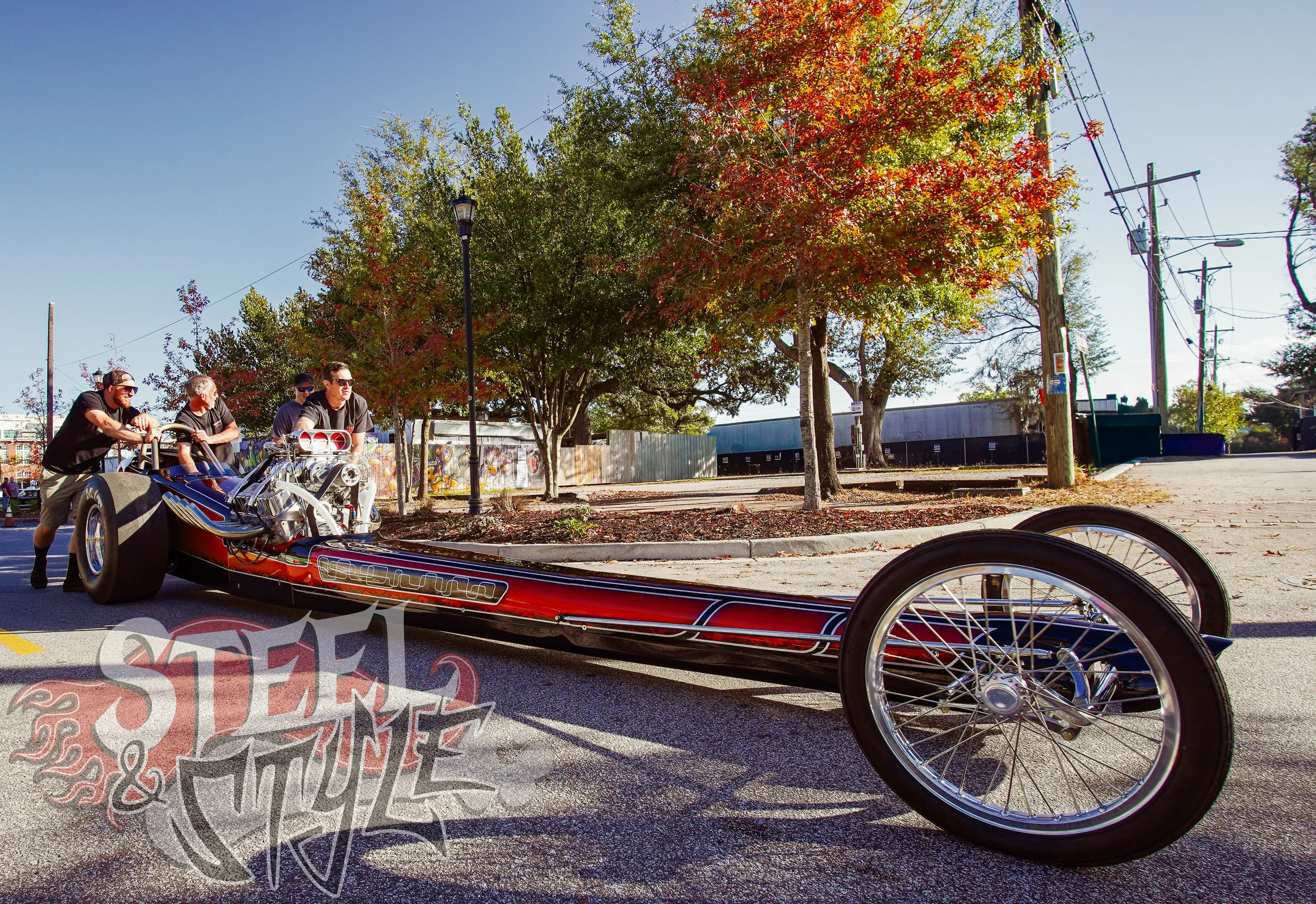 People pushing a vintage drag racing car on a city street, with colorful trees and utility poles in the background during daytime.