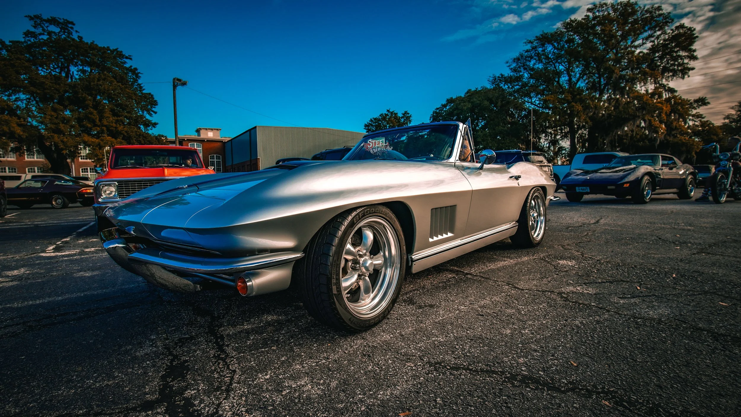 Silver vintage convertible sports car parked in a lot among other classic cars, with a blue sky and trees in the background.