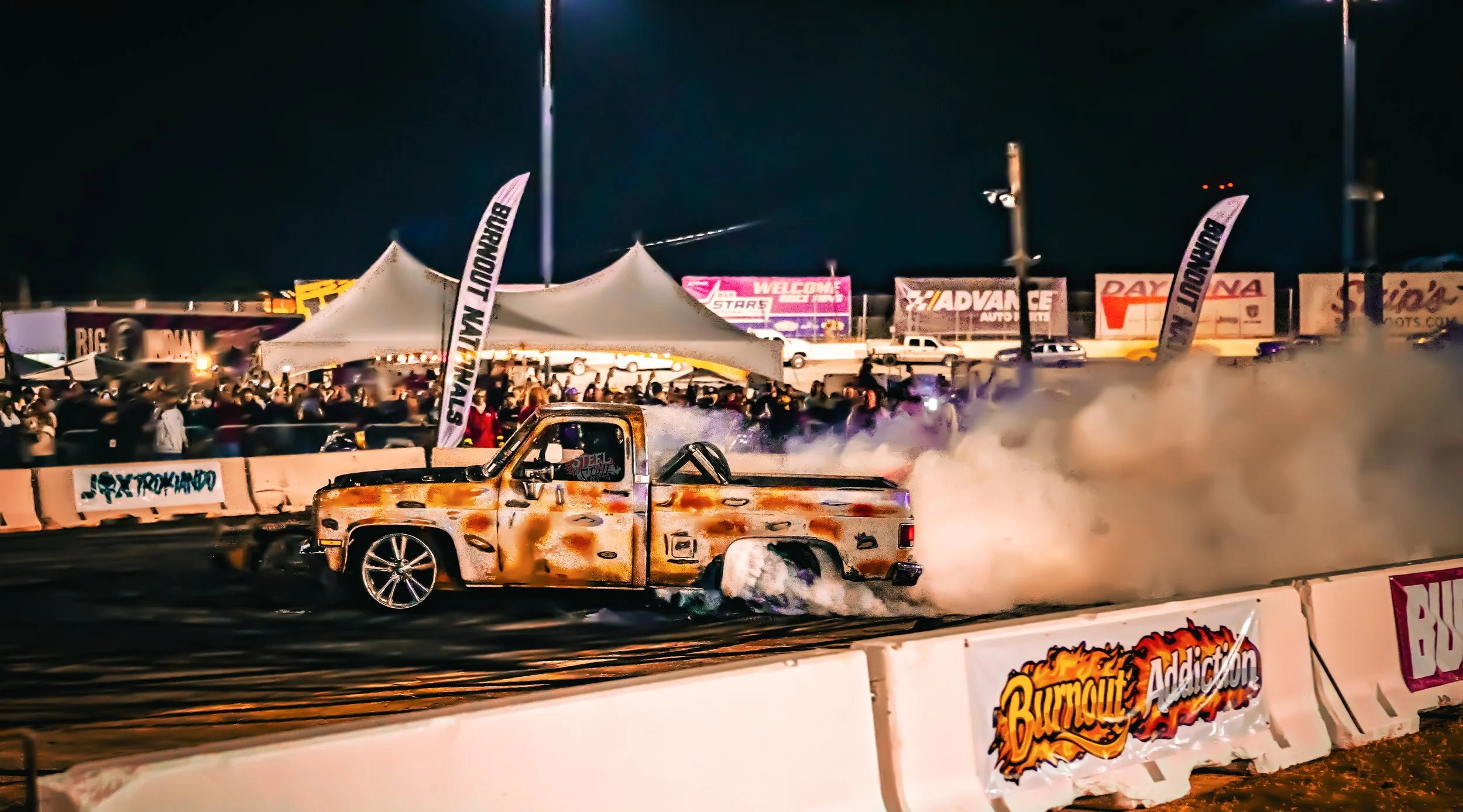A rusty pickup truck drifting at night during a burnout contest at a racing event, with smoke surrounding the vehicle and a crowd of spectators in the background.