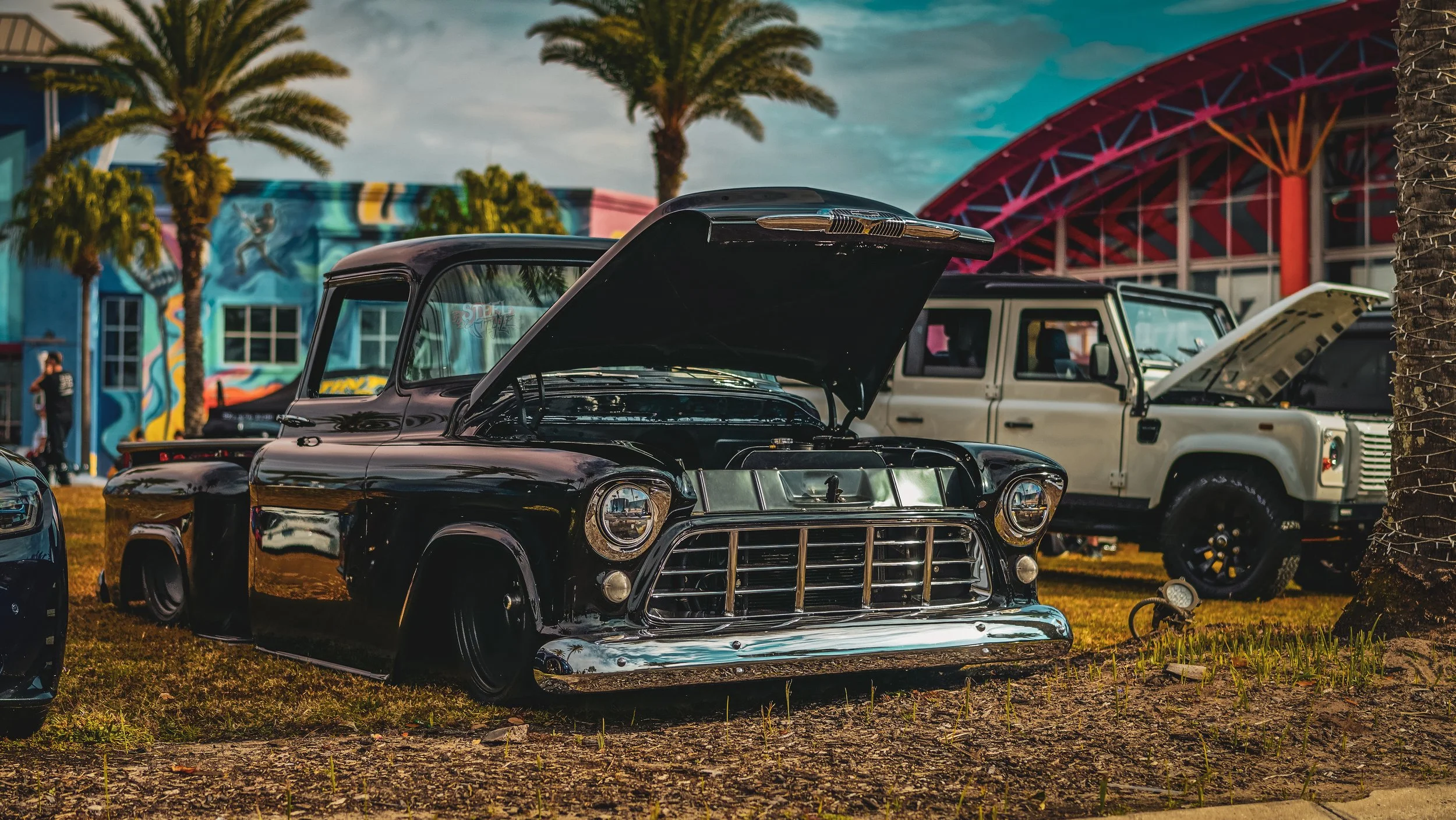 A vintage black classic car with its hood open, parked outdoors on grass with palm trees and colorful buildings in the background.