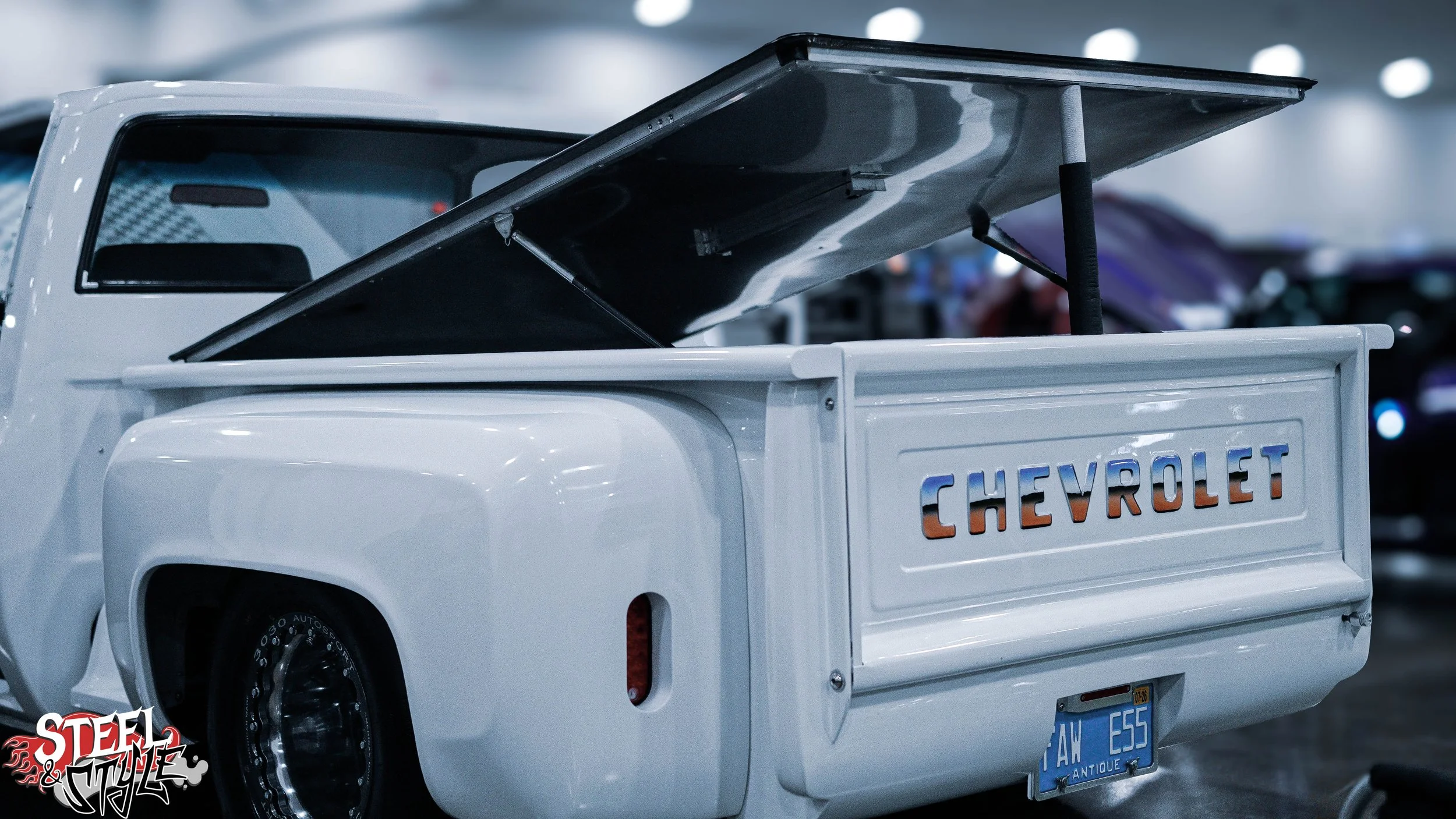 White Chevrolet pickup truck with a partially open truck bed lid at an indoor car event, with other cars and blurred lights in the background.