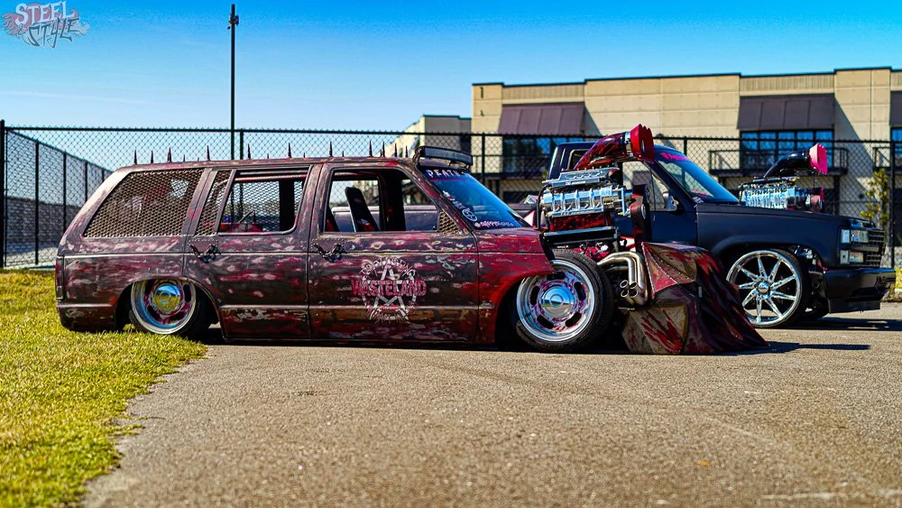 Modified lowrider car with a dark, custom paint job, lowered stance, and unique wheels, parked next to another customized truck on a paved lot with a fence and buildings in the background.