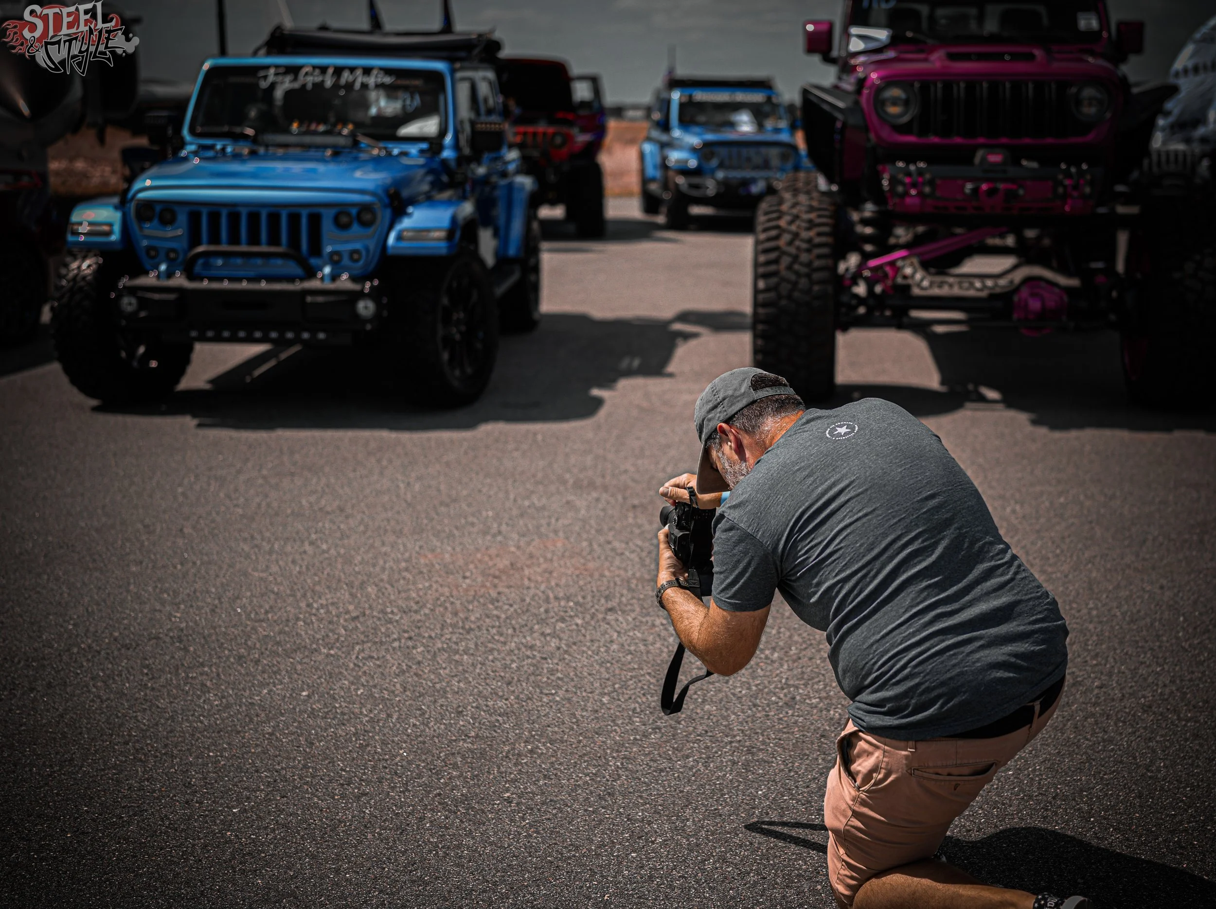 Photographer crouching on paved ground taking a picture of colorful off-road vehicles parked in the background.