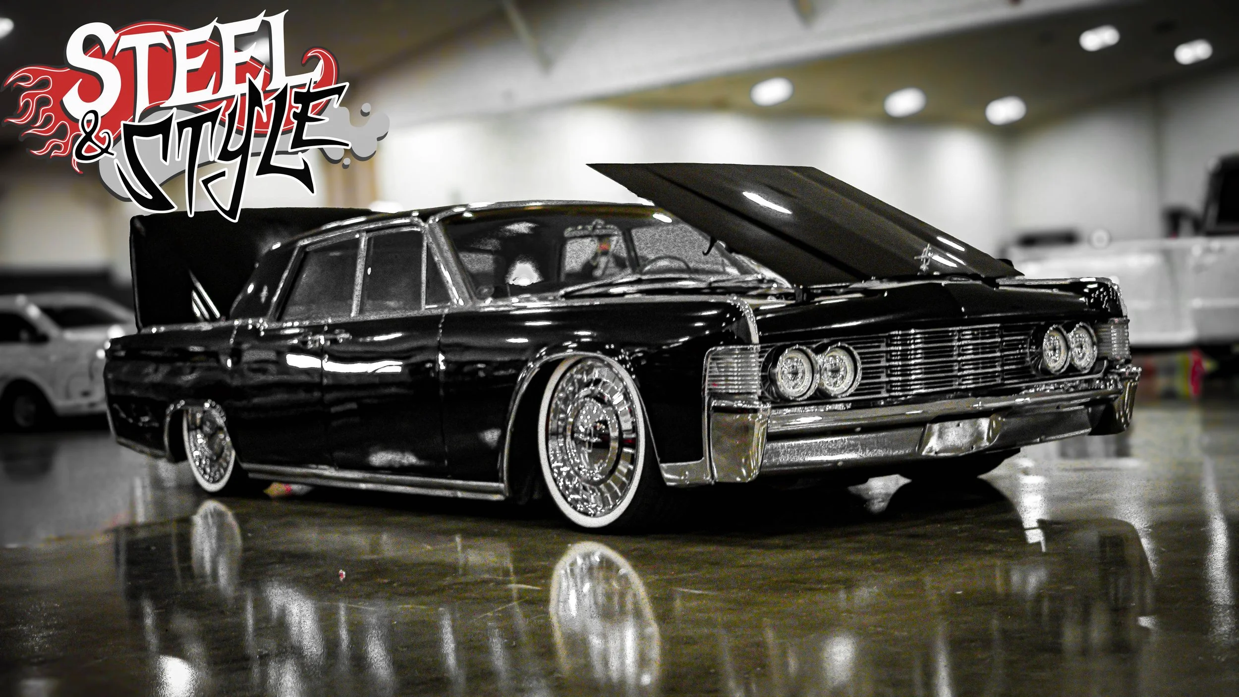 A vintage black lowrider car with chrome accents and white-walled tires, displayed indoors on a reflective floor. A colorful 'Steel & Style' logo is in the upper left corner.