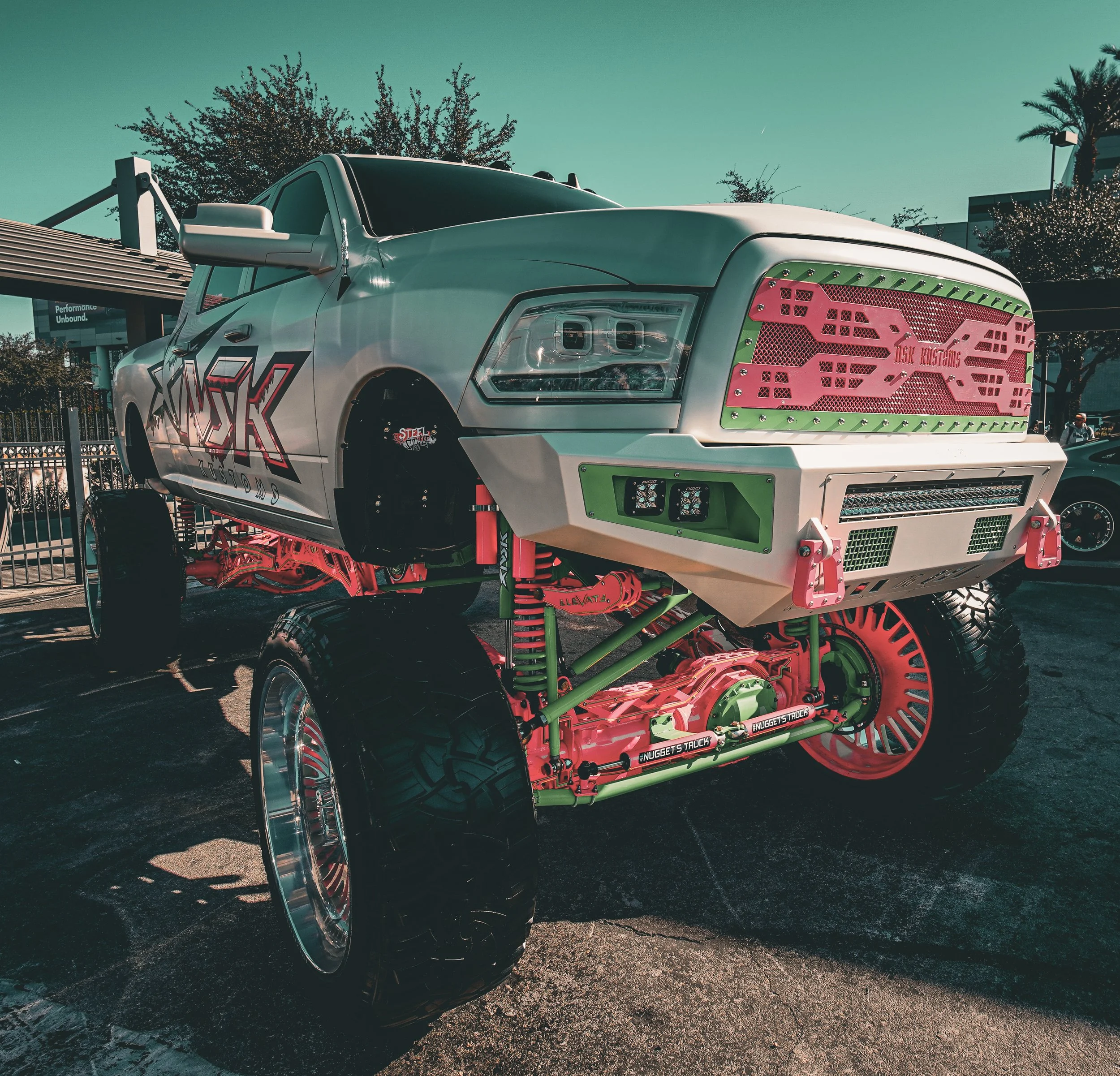A modified lifted truck with oversized tires, green and pink accents, custom grille, and exposed suspension components.