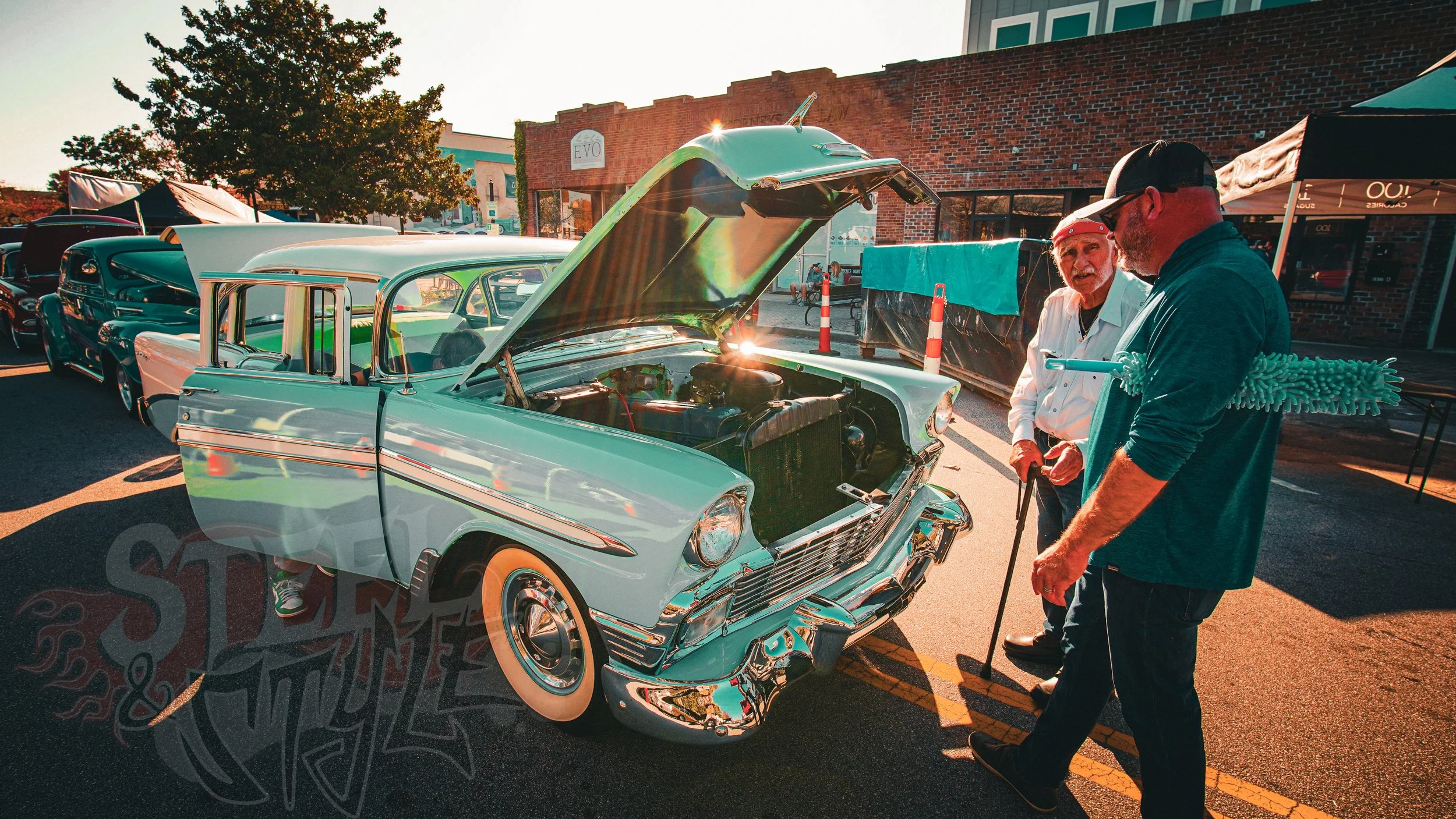 Two men standing near a white vintage car with its hood open at a car show, with other classic cars parked nearby amidst a sunny outdoor setting.