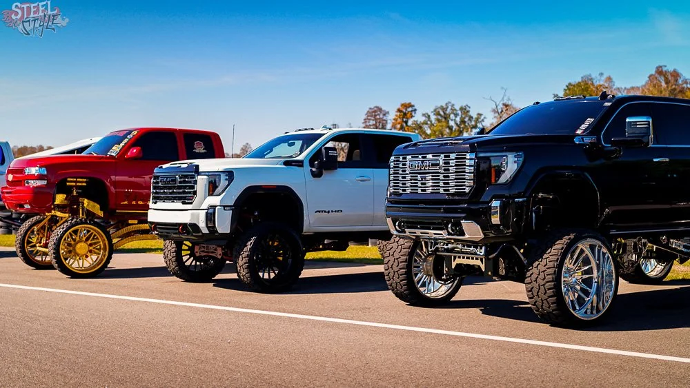 Three lifted trucks and a vintage tractor on display at a car show. The trucks are a red, white, and black vehicle, each with unique custom wheels and suspension.