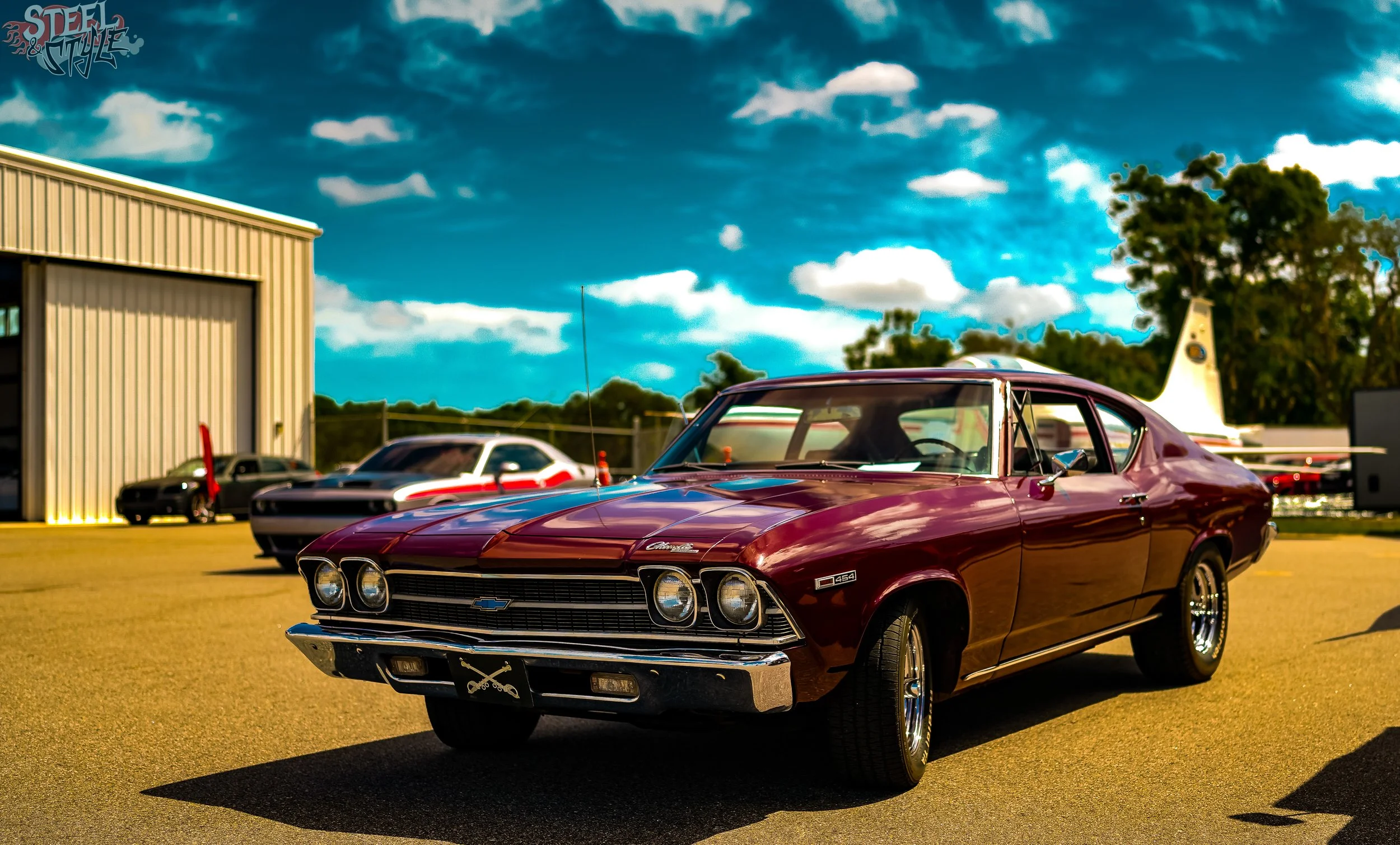 A vintage red Chevrolet Chevelle SS is parked outdoors on a sunny day at a classic car show, with other classic cars and a large industrial building in the background.