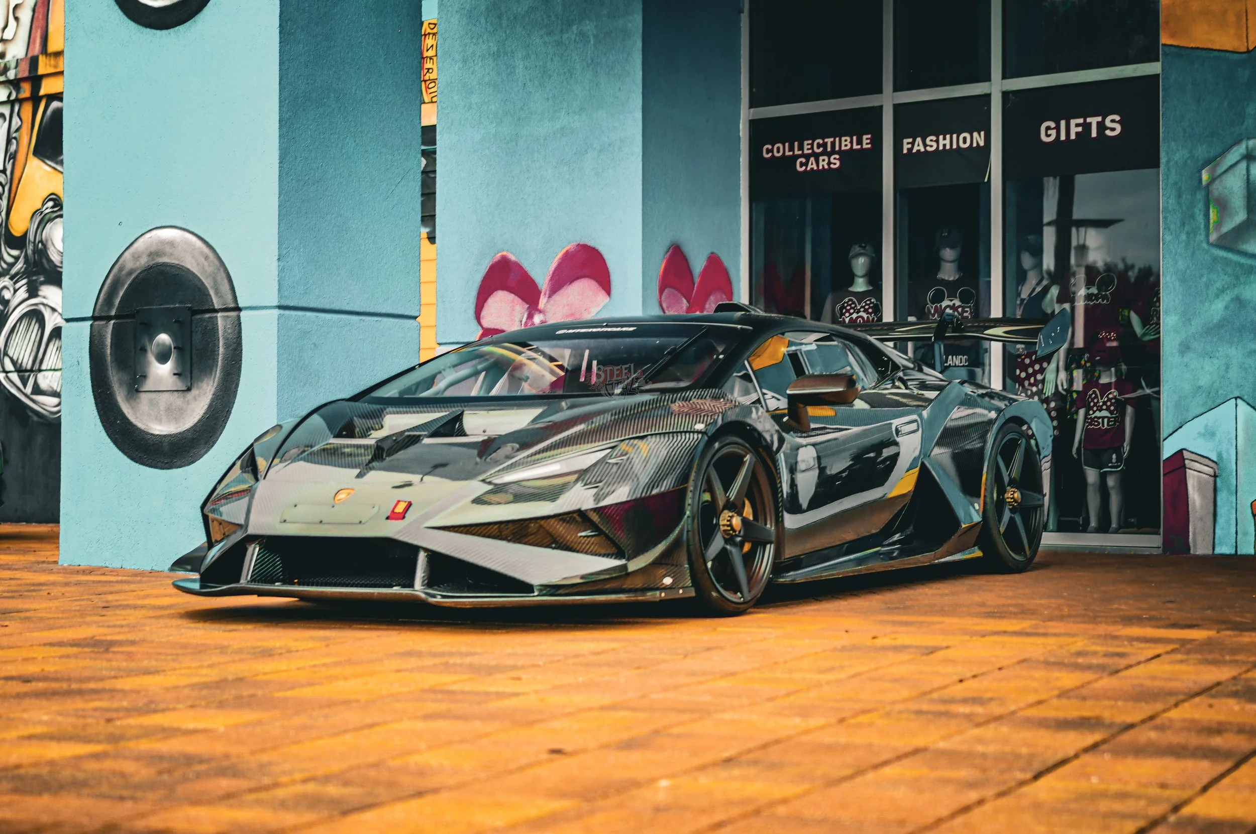 A sleek black and yellow sports car parked on a brick-paved street in front of a store with mannequins and colorful graffiti art.