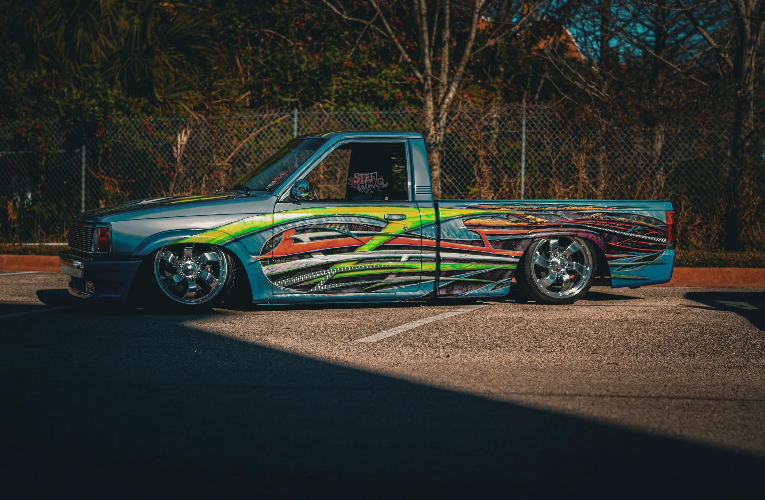 A lowrider pickup truck with a colorful, intricate flame and wave graphic design on its body, parked in a lot during daytime.
