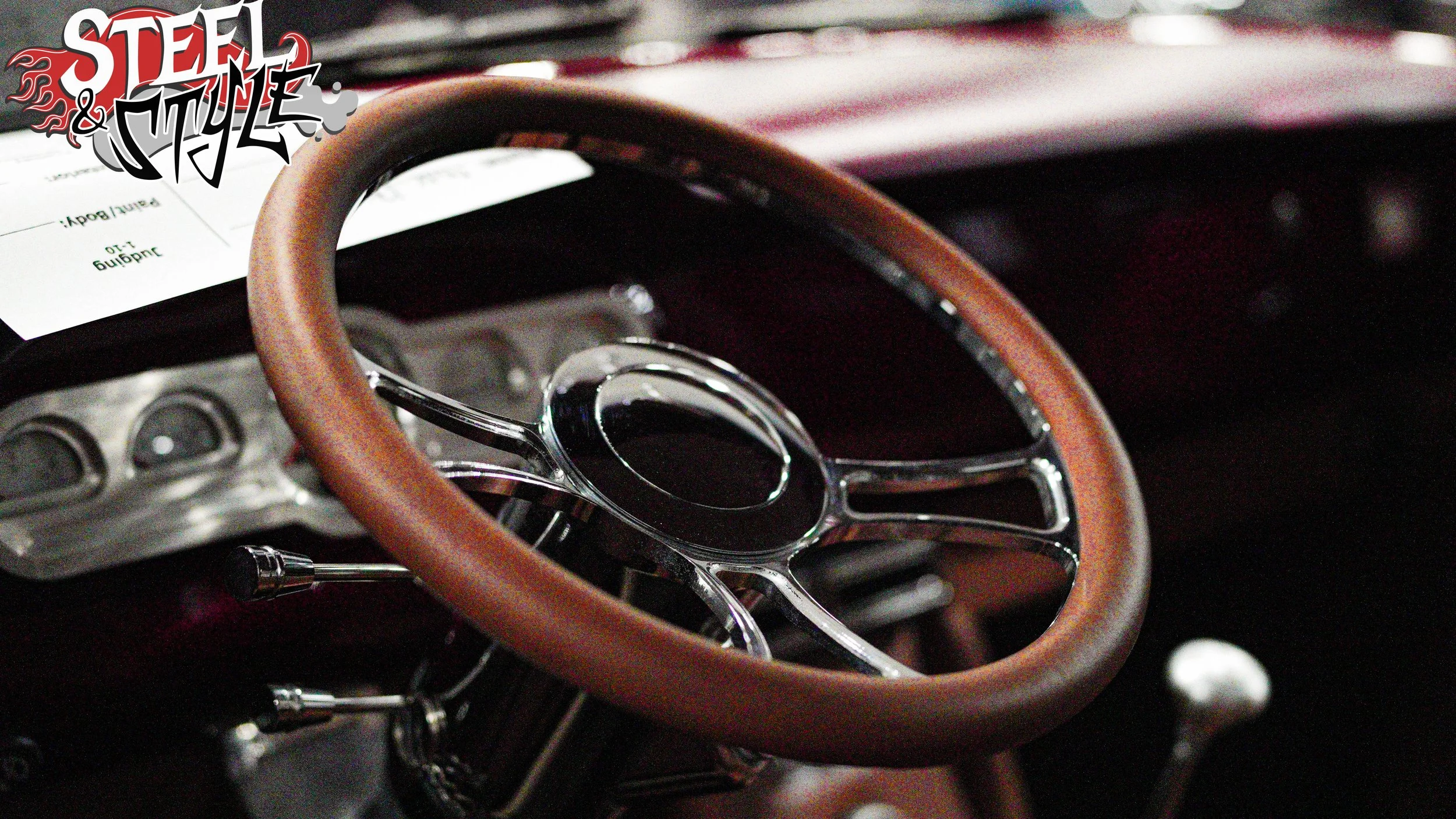 Close-up of a vintage car's steering wheel with a brown leather grip and metal spokes, interior dashboard visible in the background.