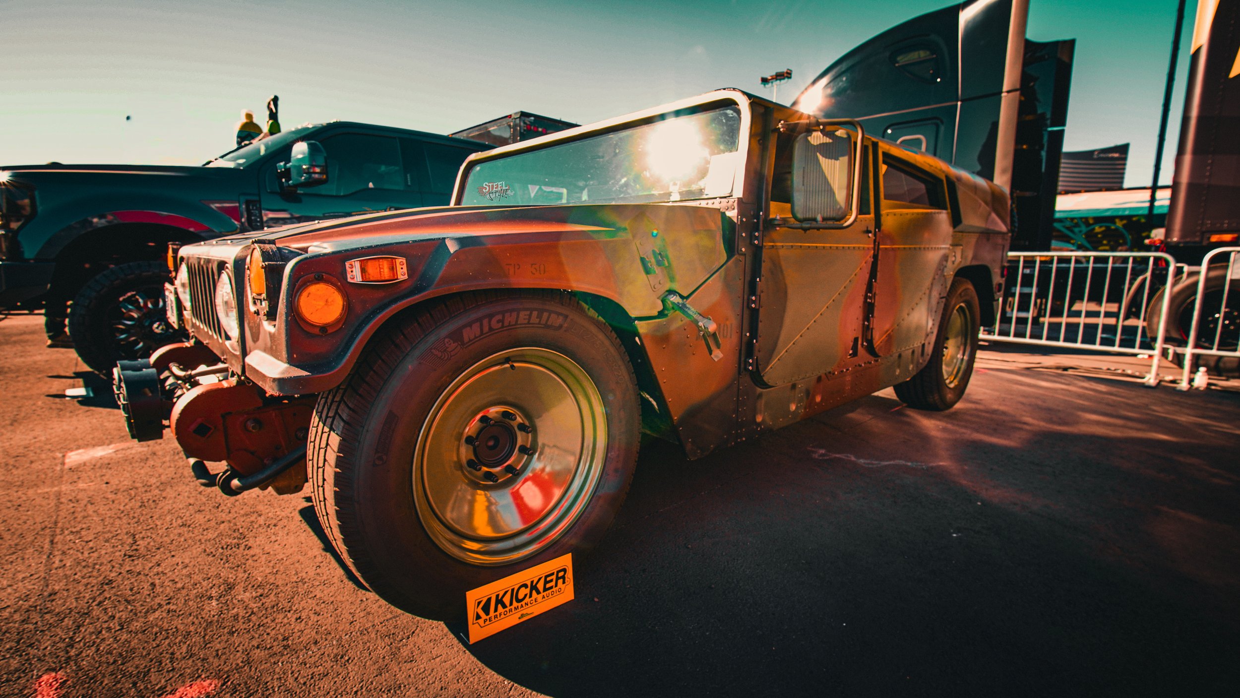 A vintage military-style vehicle with a rugged, weathered look on display at a car show, with a sign that reads 'KICKER' in front of the front wheel.