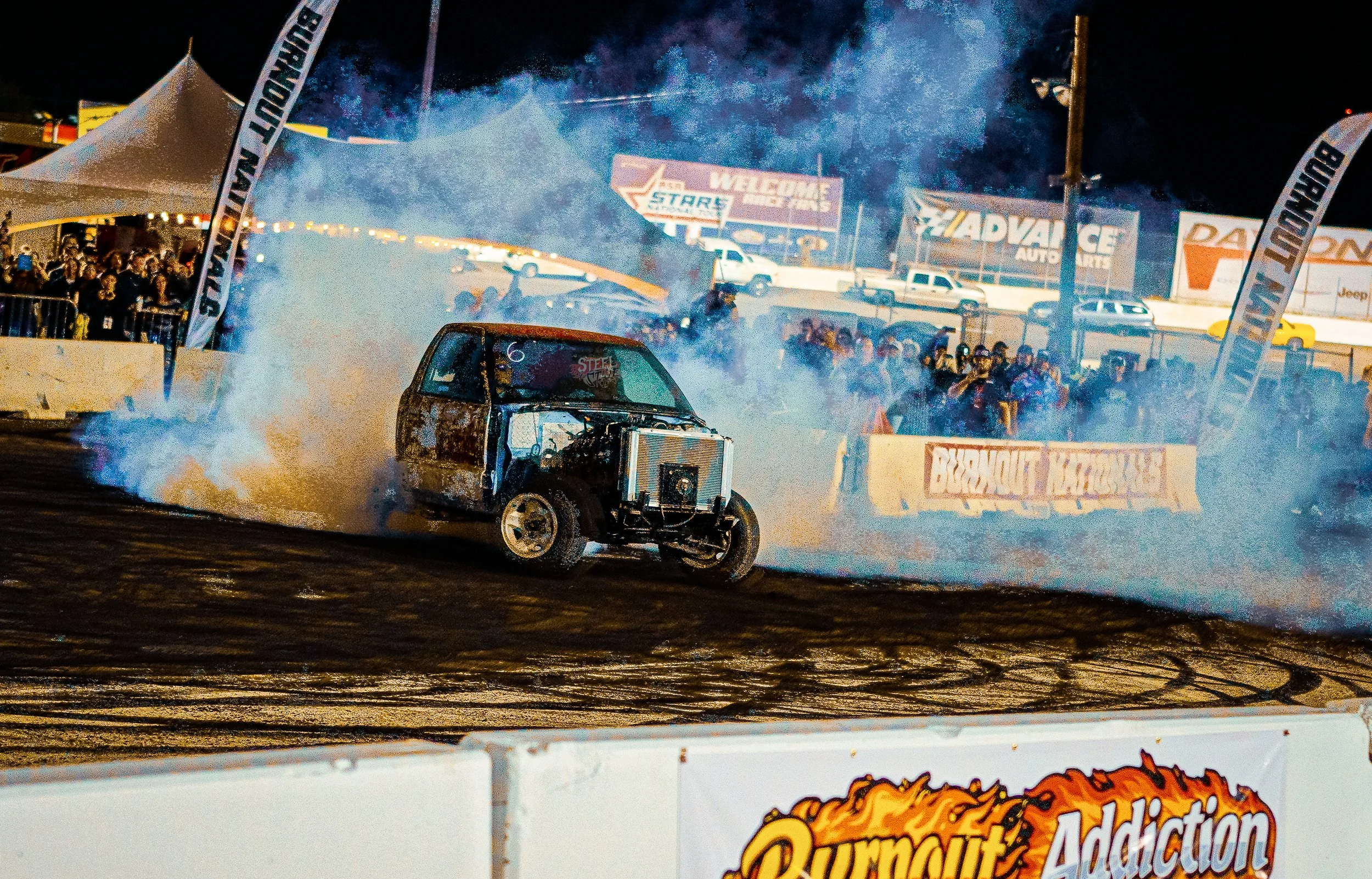 Nighttime dirt track car race with a damaged vehicle emitting smoke on the track, spectators in the background, and racing banners and advertisements.