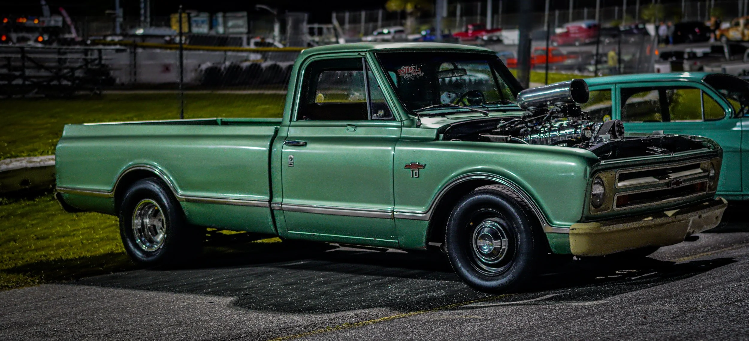 A vintage green Chevrolet pickup truck with an exposed engine is parked outdoors at night, next to a teal-colored car, with a car park and fence in the background.