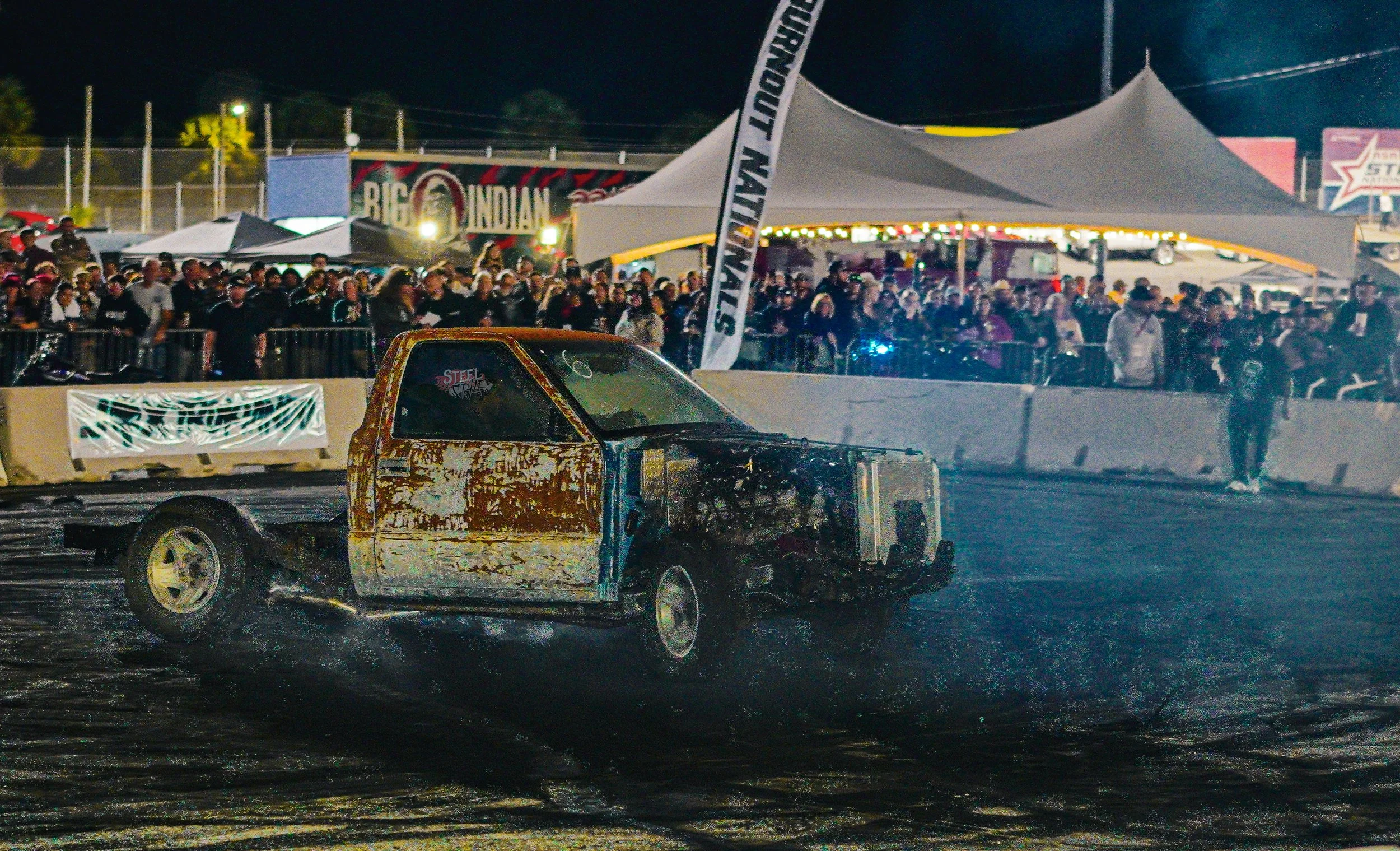 A rusted, damaged car on a racetrack during a night event with a crowd of spectators and event tents in the background.