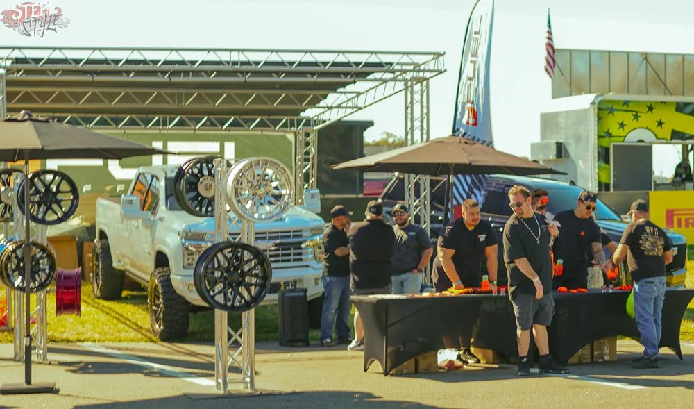 People gathered at an outdoor automotive event, with trucks, wheels displayed on stands, and food tables under umbrellas.