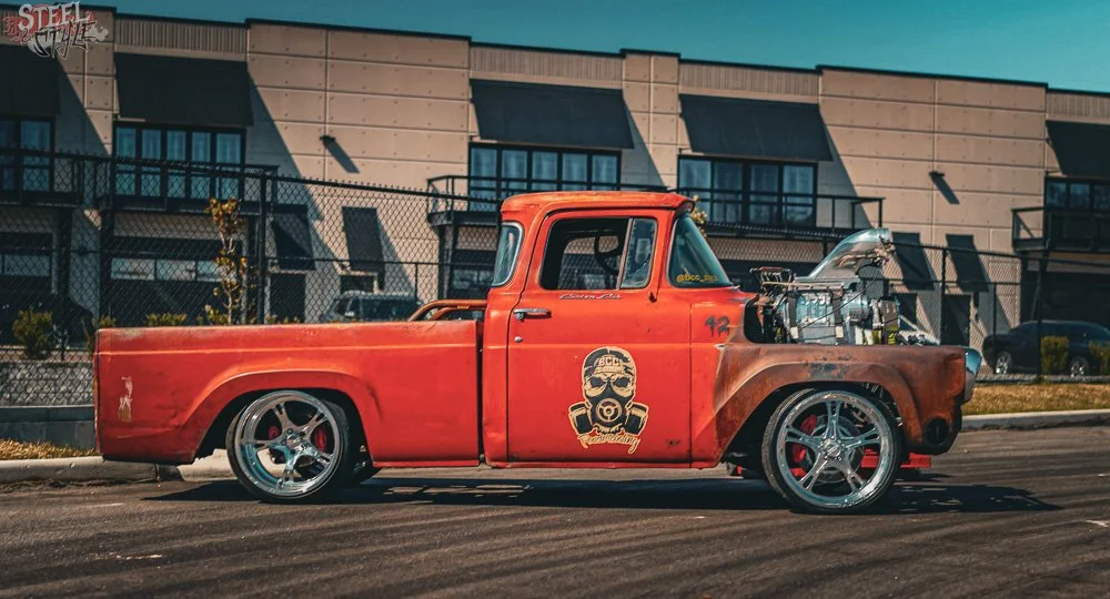 Red vintage pickup truck with large chrome wheels, custom graphics on the door, and a visible engine in the open hood, parked on the street in front of a modern residential building.