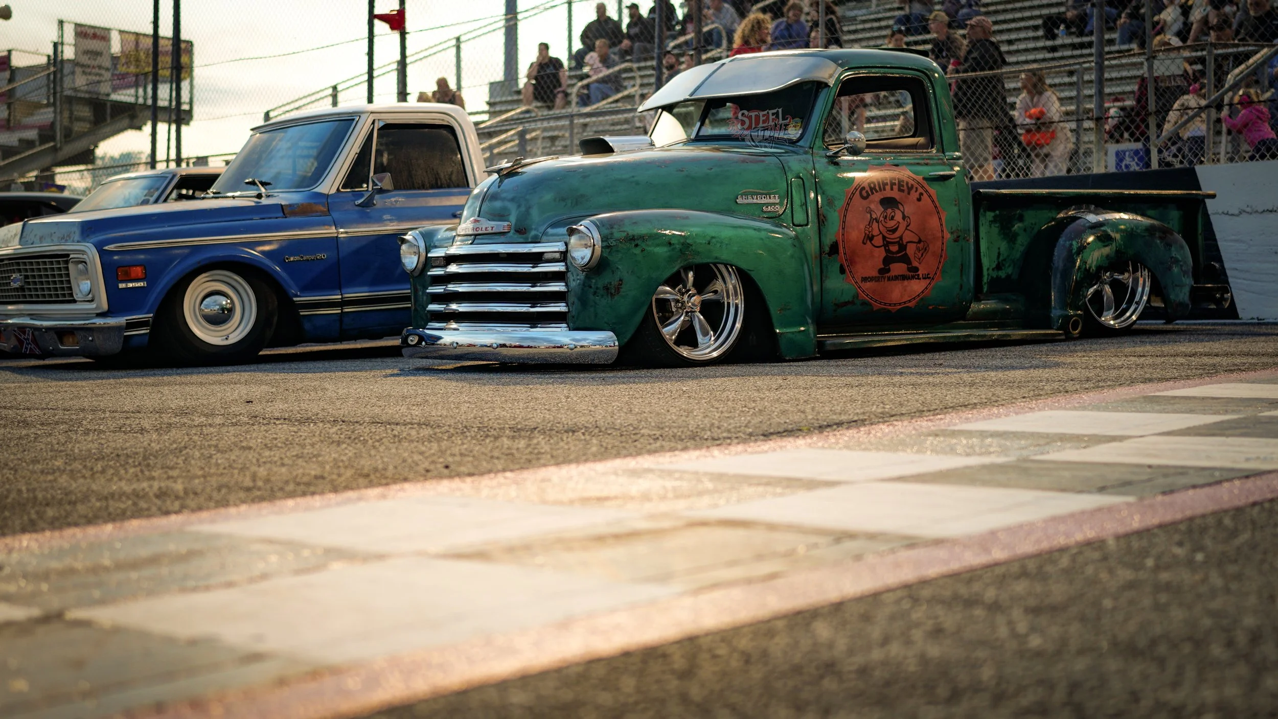 Vintage modified trucks at a race track with spectators in the stands.
